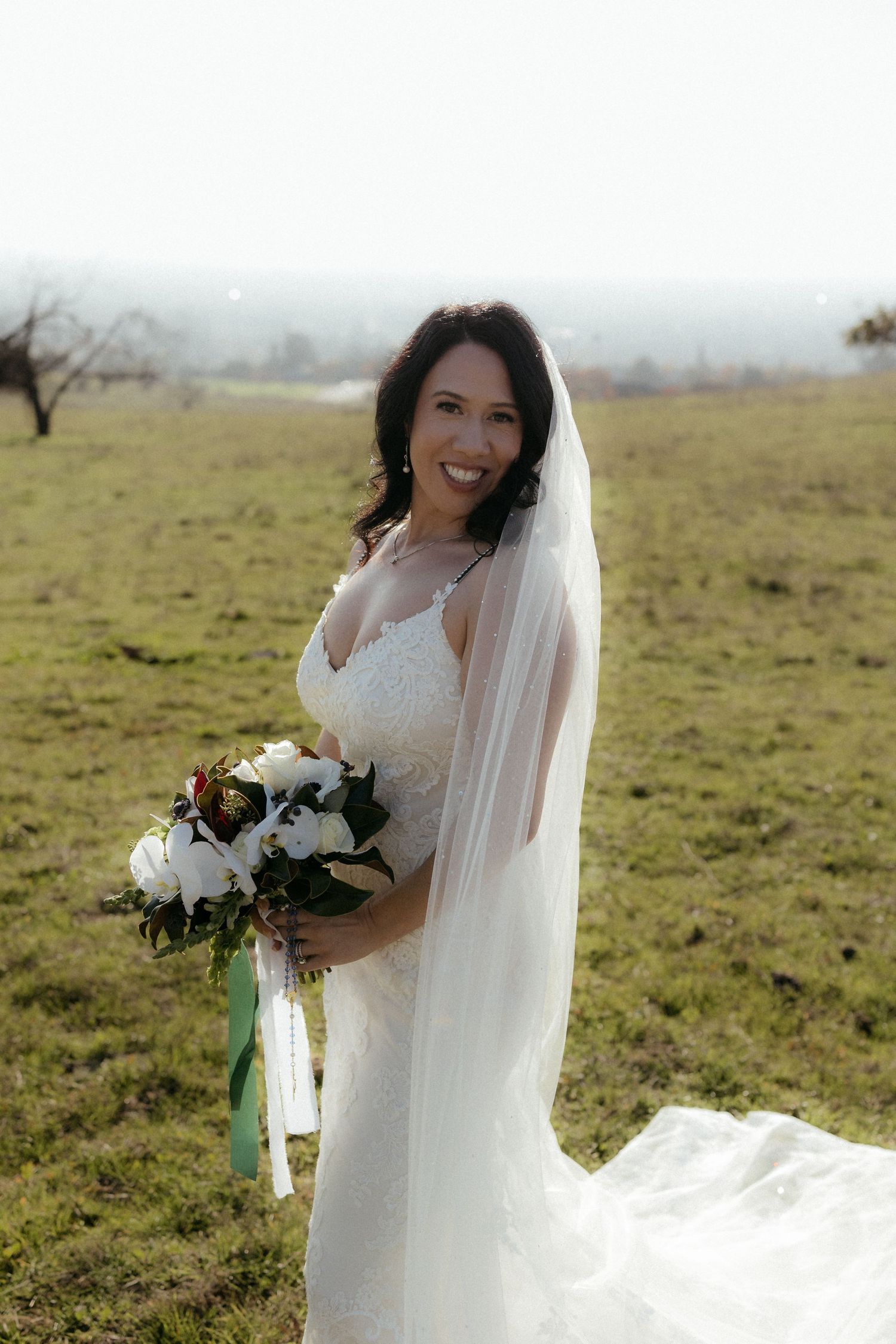 A bride in a white lace gown holds a bouquet of white flowers while standing in a sunlit meadow.