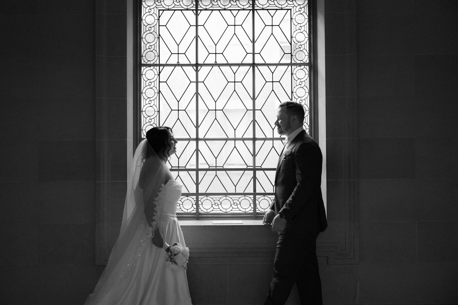A silhouetted couple stands by a geometric patterned window in dramatic black and white photography.