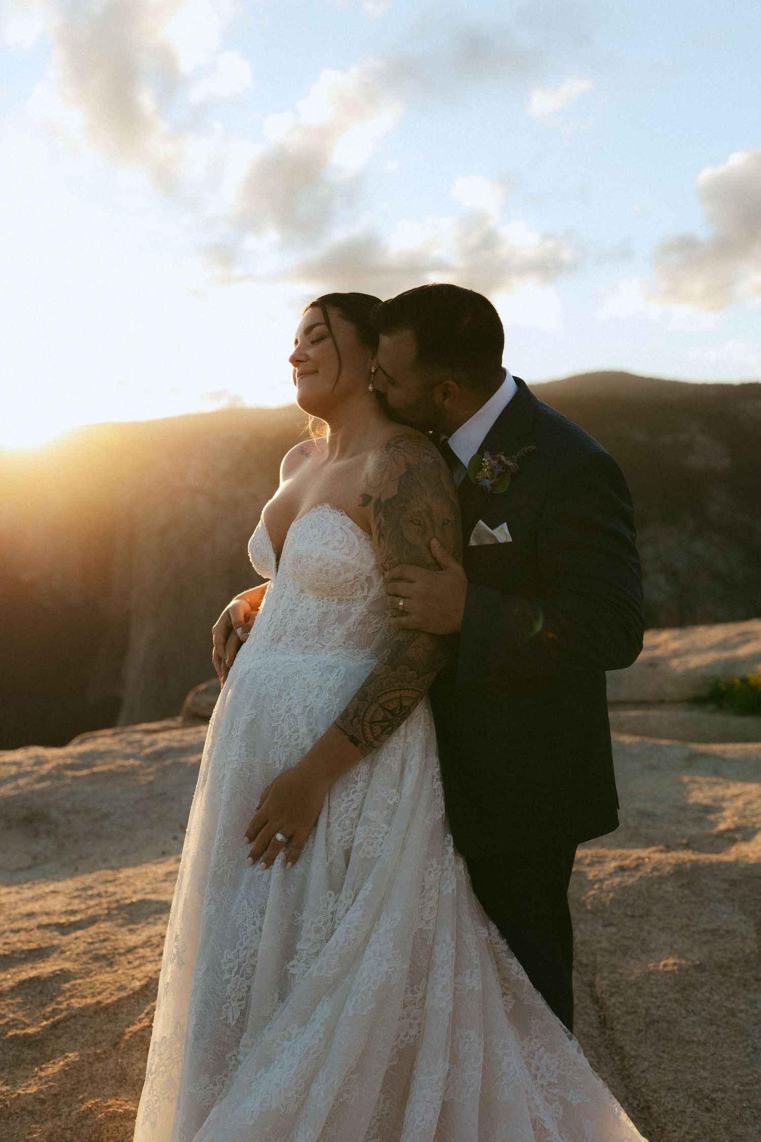 Groom with tattoos kisses bride in lace wedding dress at sunset on rocky Yosemite mountain overlook.