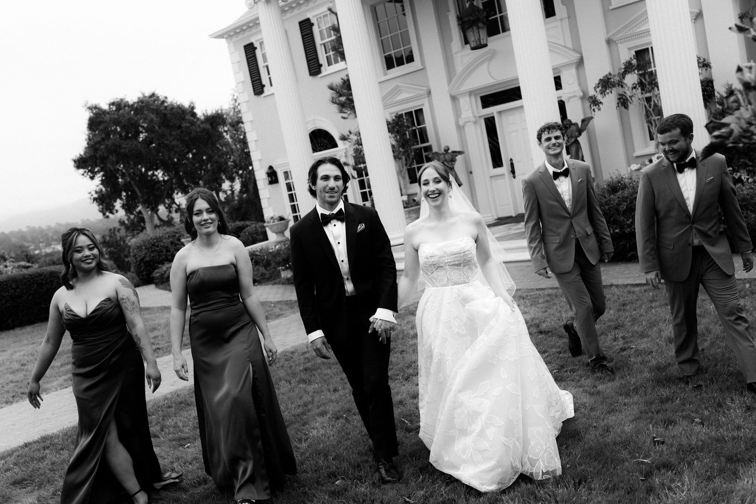 Black and white wedding photo of bride and groom walking with bridal party outside elegant white mansion.