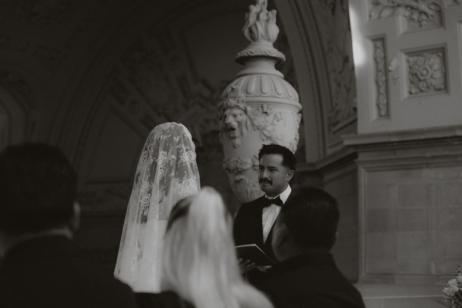 Black and white wedding ceremony photo with bride in veil and groom holding book before ornate stone column.