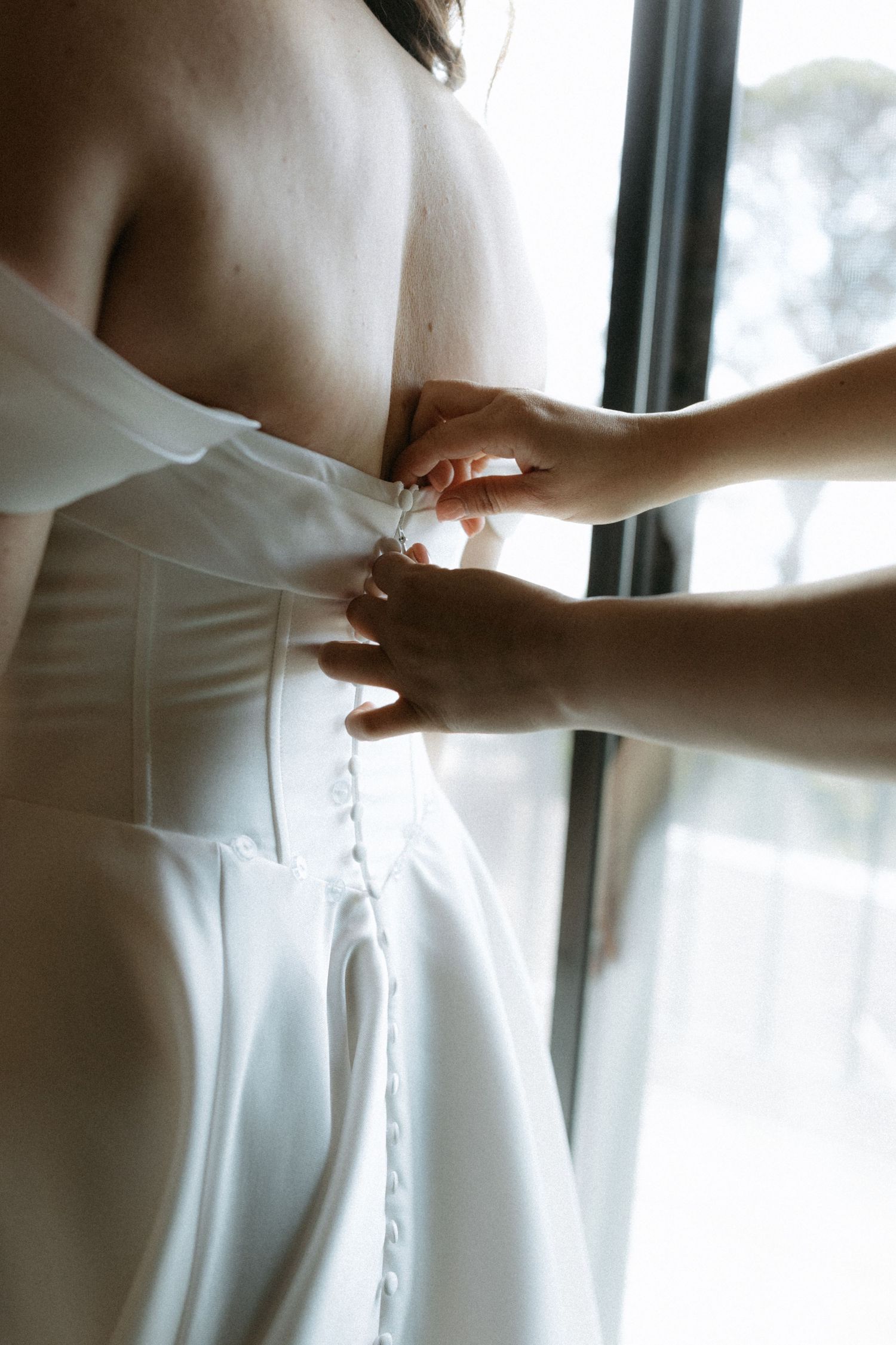 Close-up of a bridesmaid fastening the back buttons of a white wedding dress by a bright window.