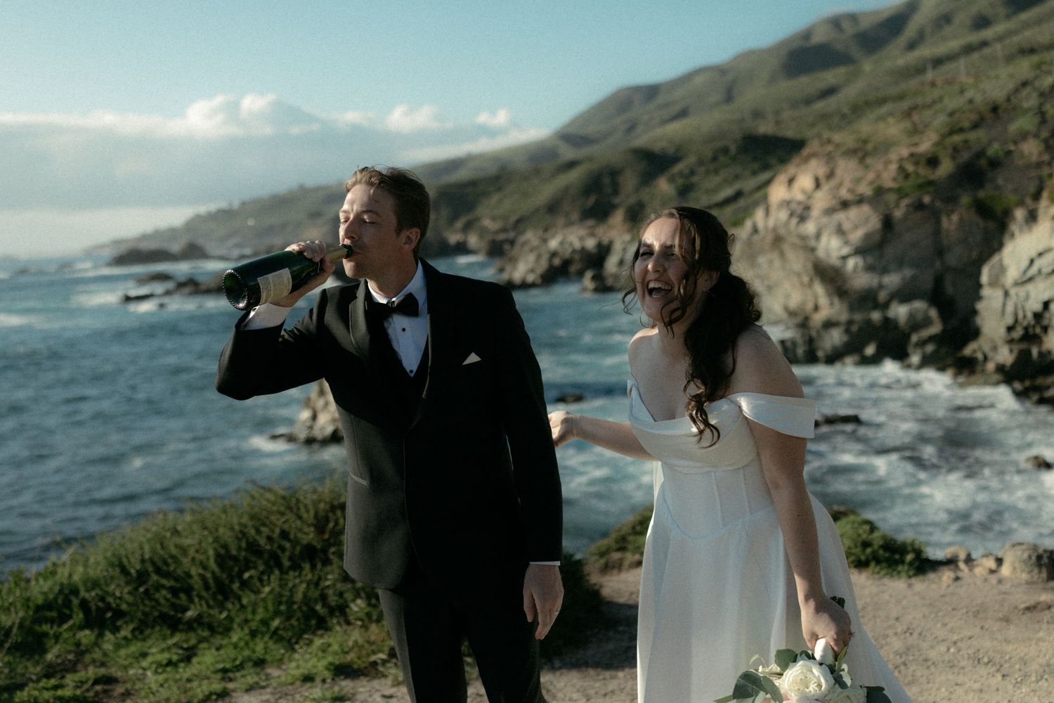 Bride and groom stand on a rocky Big Sur coastline, groom drinking from a flask in a black tuxedo, bride in white gown.