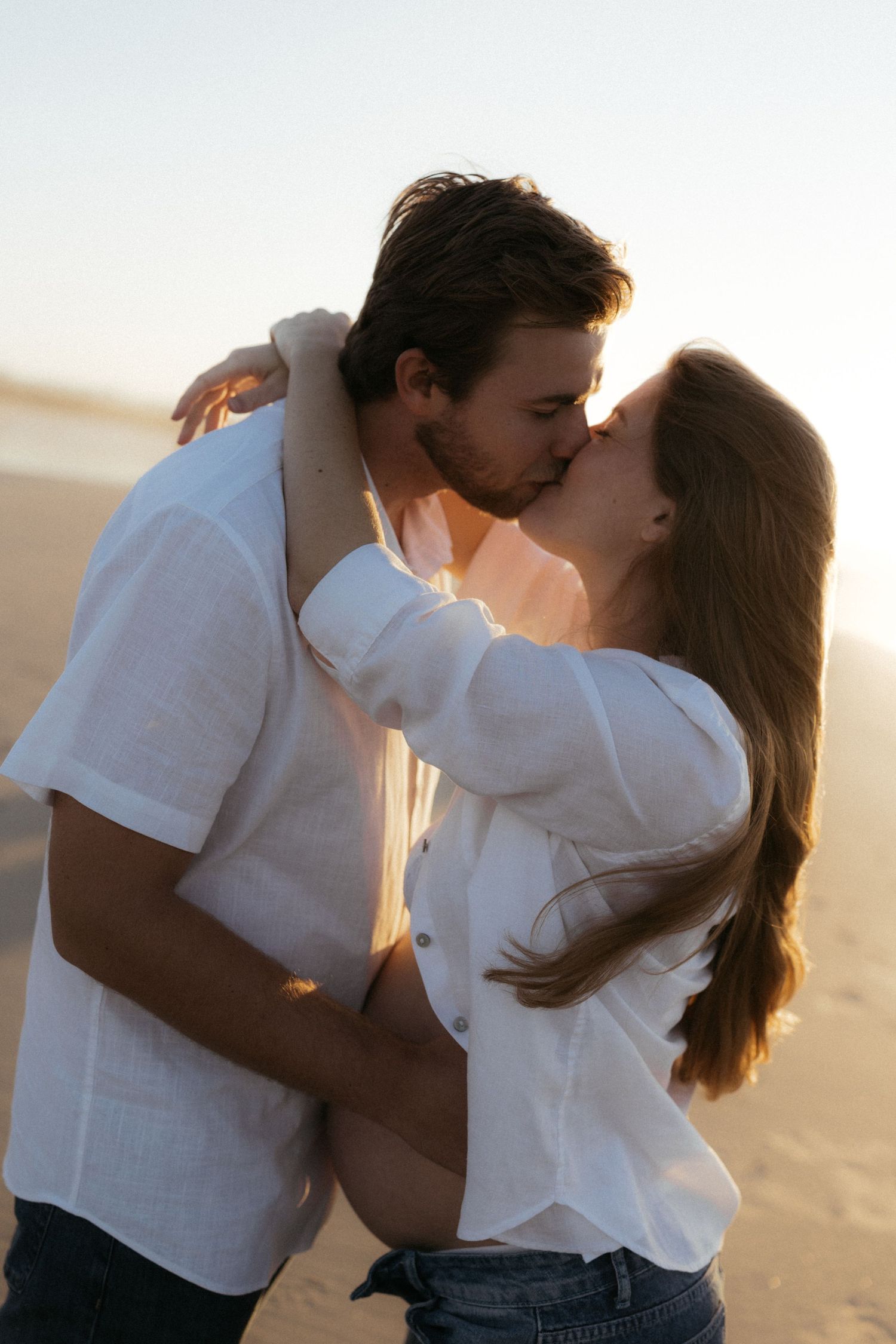 Romantic couple sharing a kiss on a sandy beach during golden sunset, both wearing white shirts.
