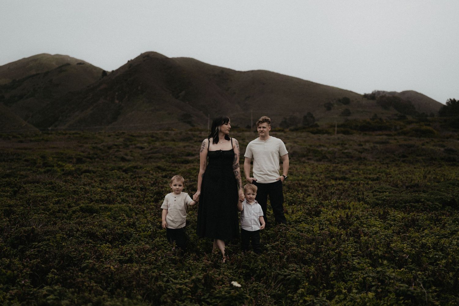 Family of four standing in a lush green field with rolling hills in the background on a cloudy day.