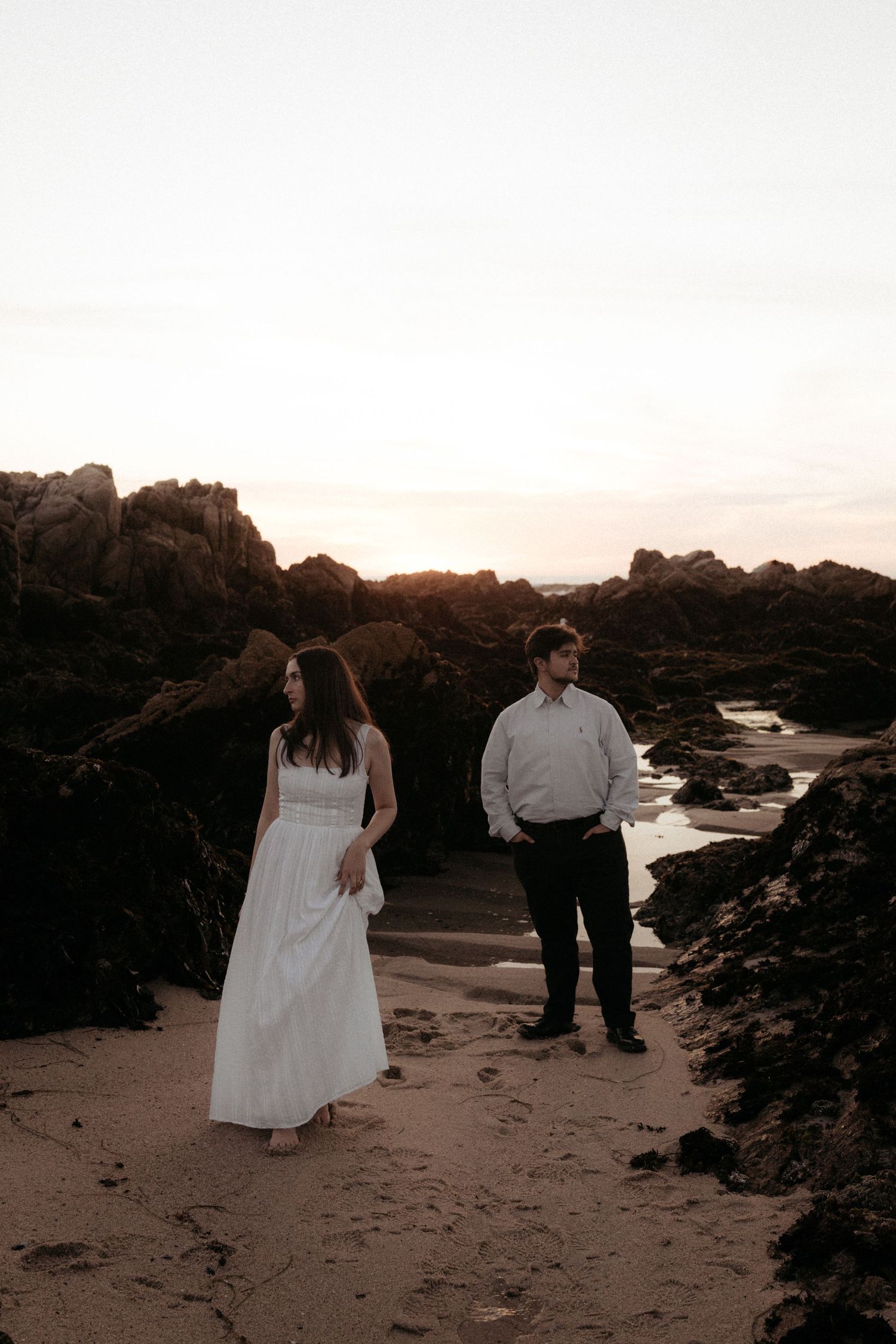 A couple walks along a sandy beach at sunset surrounded by dark rocky cliffs.