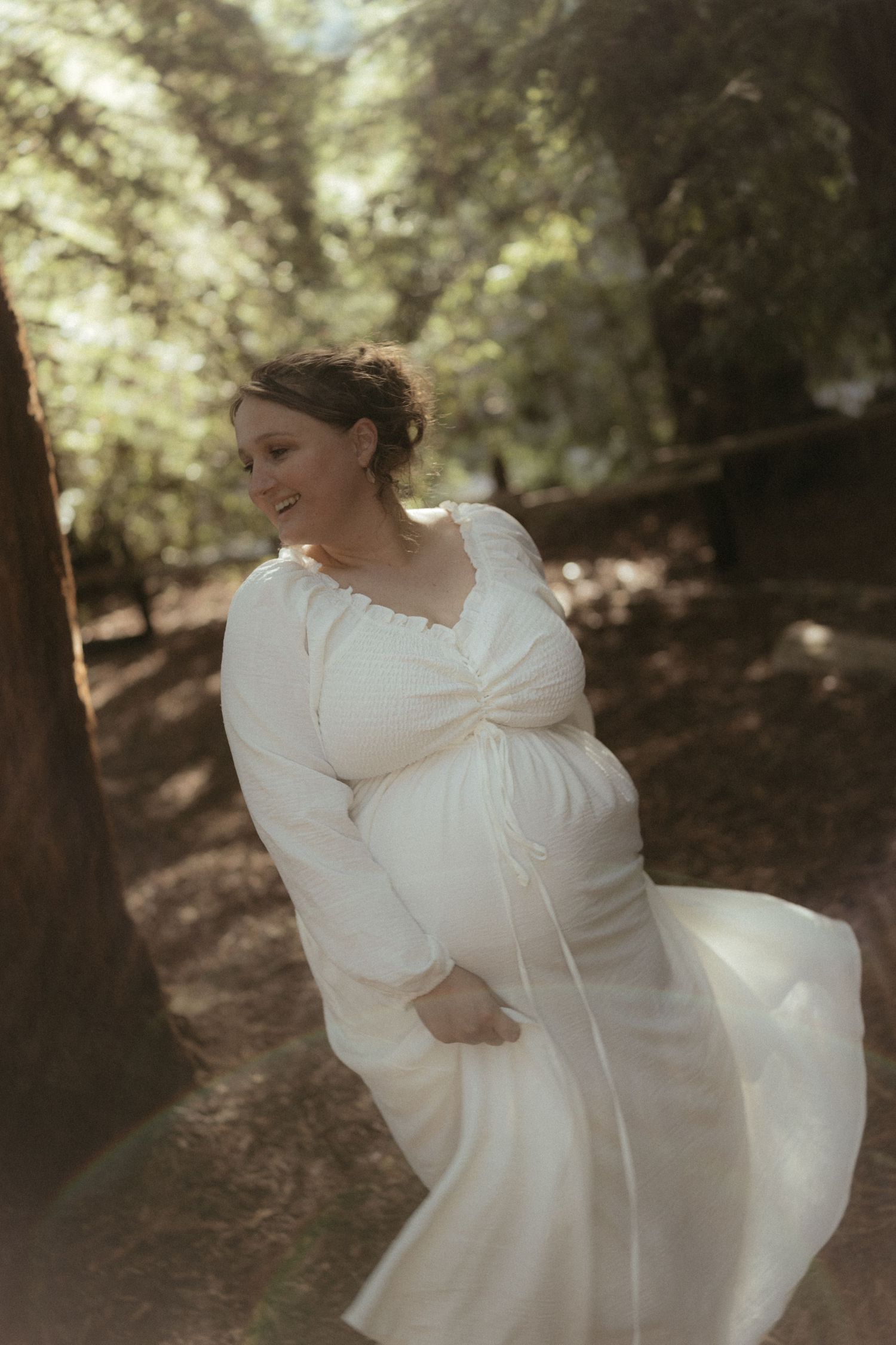 A person in a white dress twirls playfully in a sun-dappled forest setting.