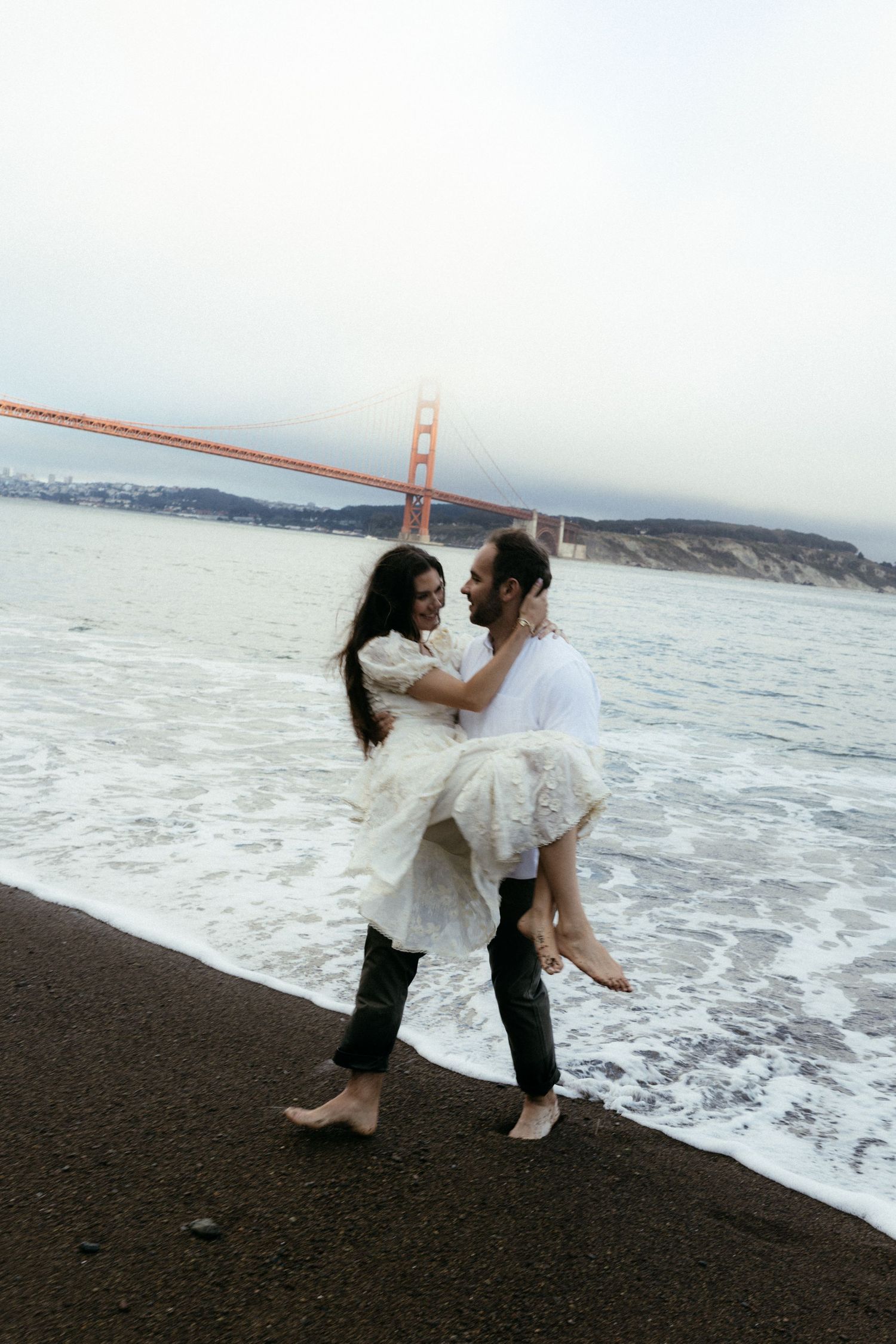 Couple sharing romantic moment on beach near Golden Gate Bridge, man carrying woman in white dress at water's edge.