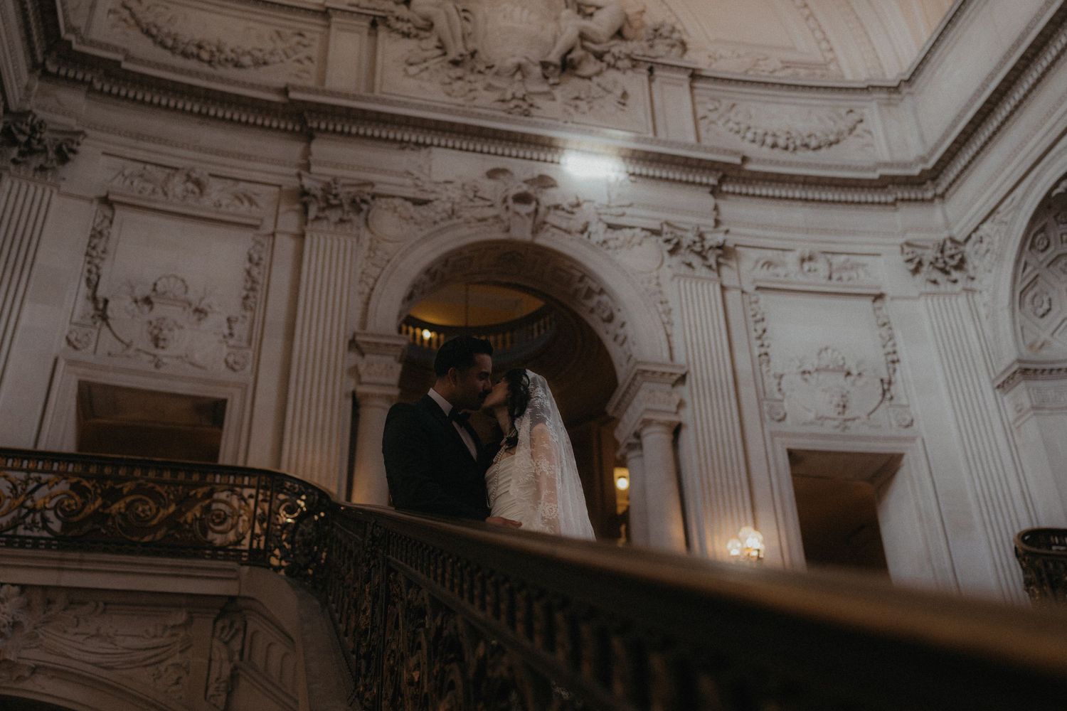 Bride and groom share an intimate moment on an ornate balcony in a grand white baroque-style venue.
