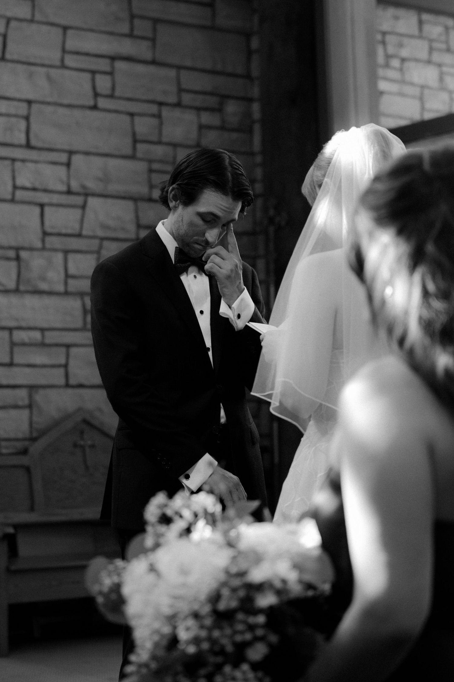 Emotional groom wiping tears in black and white wedding ceremony photo with bride in veil.