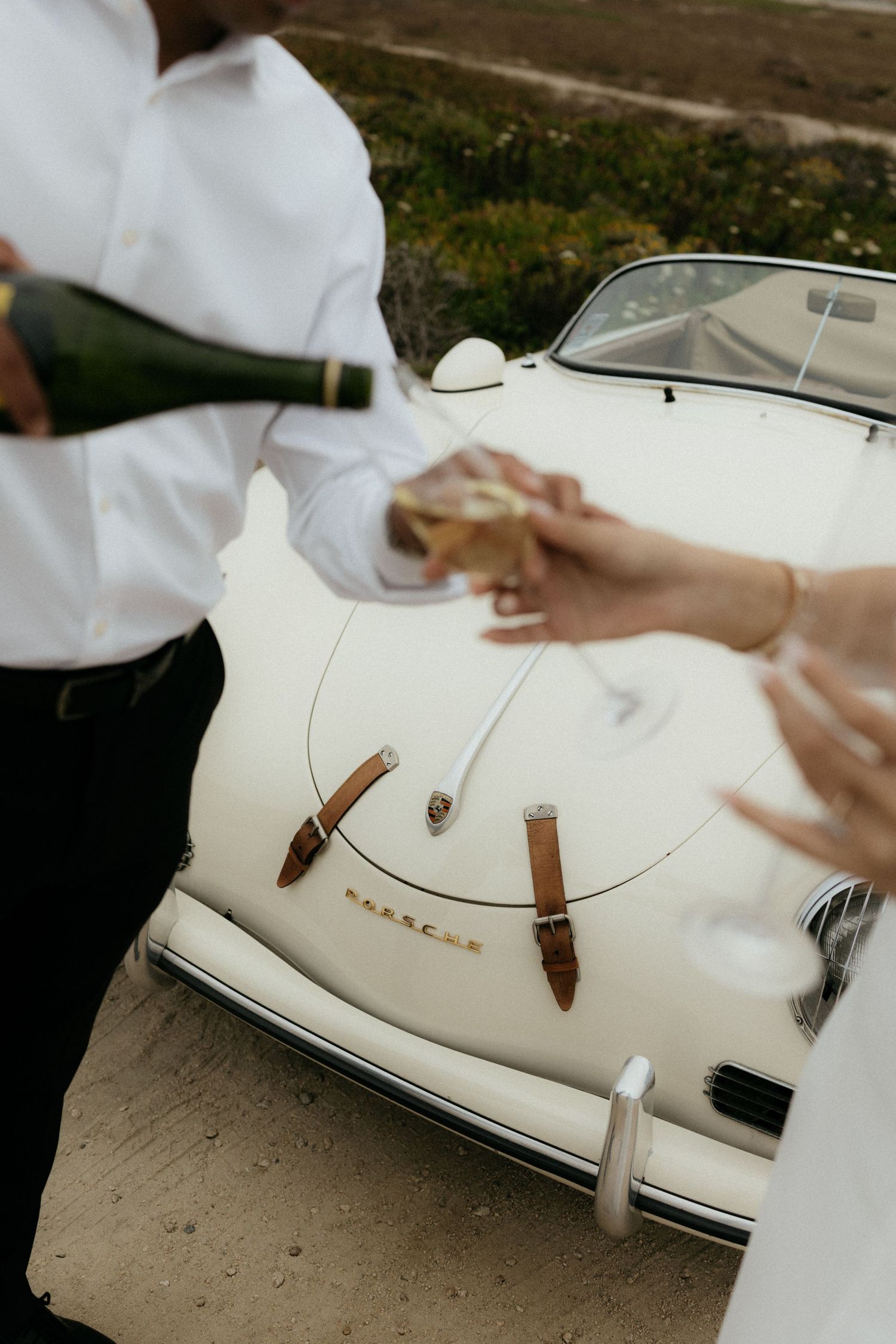 Couple pouring champagne beside a classic white Porsche convertible during outdoor wedding celebration.