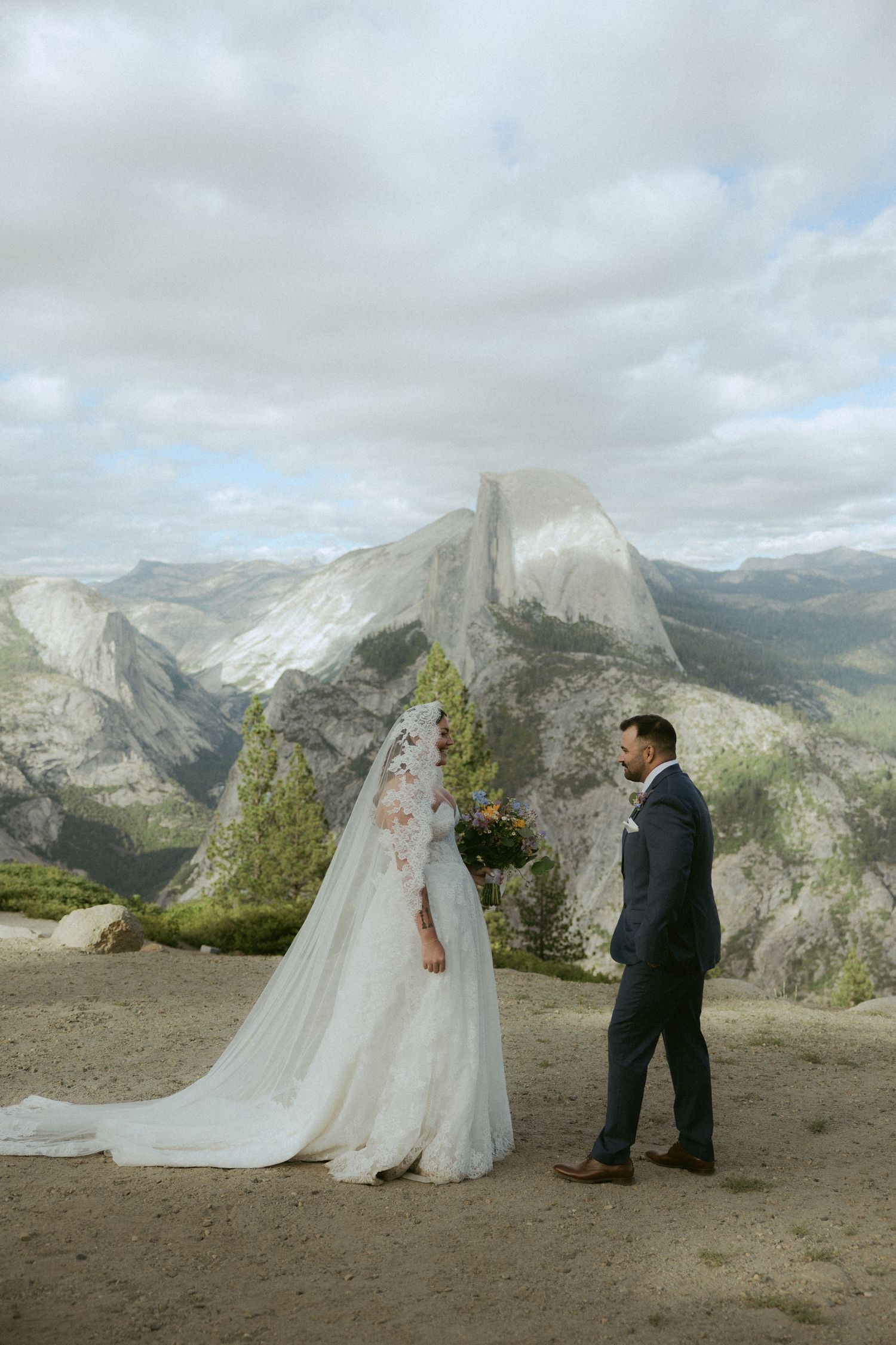 Bride and groom stand at Glacier Point with Half Dome backdrop in Yosemite.