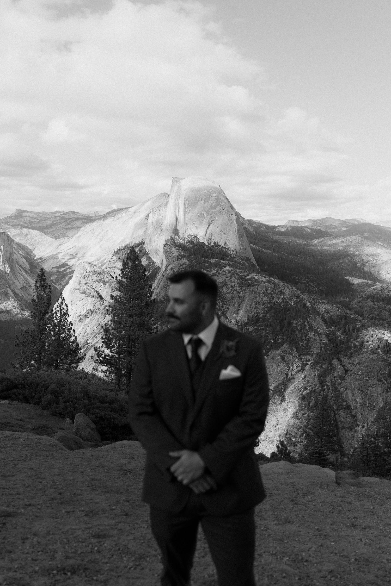 Groom in dark suit stands before Half Dome at Yosemite in black and white wedding portrait.