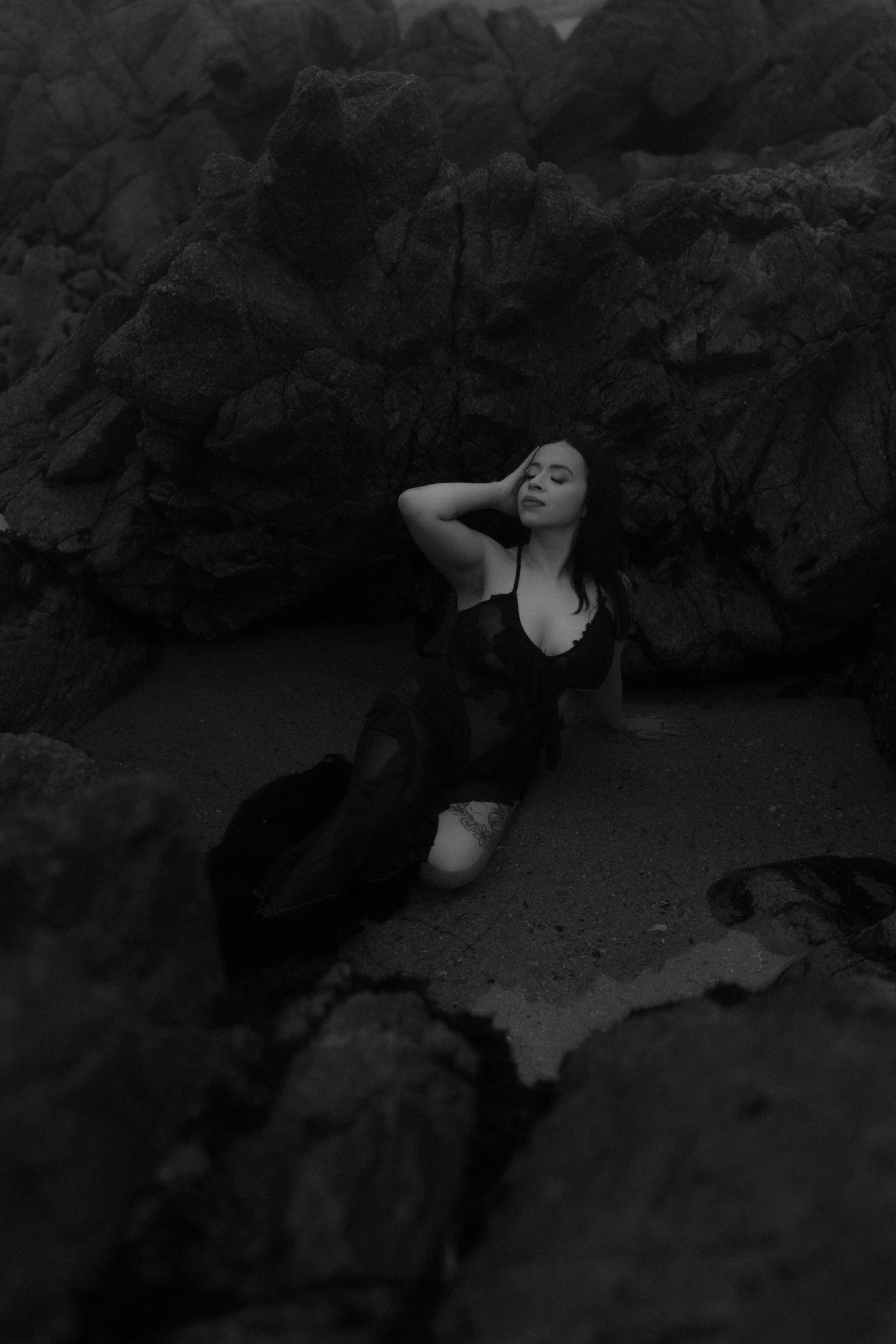 A person in black swimwear reclines dramatically against dark rocky cliffs in a moody black and white photograph.