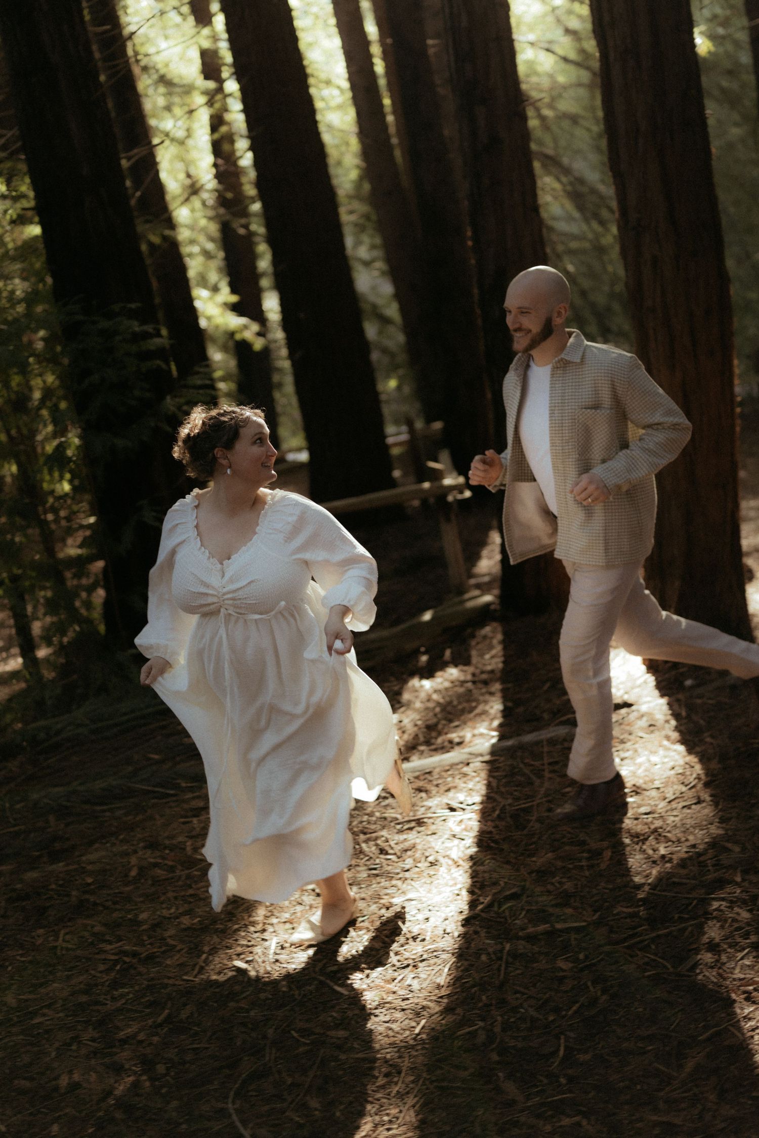Dancing figures in flowing white clothing move gracefully between towering forest trees in dappled sunlight.