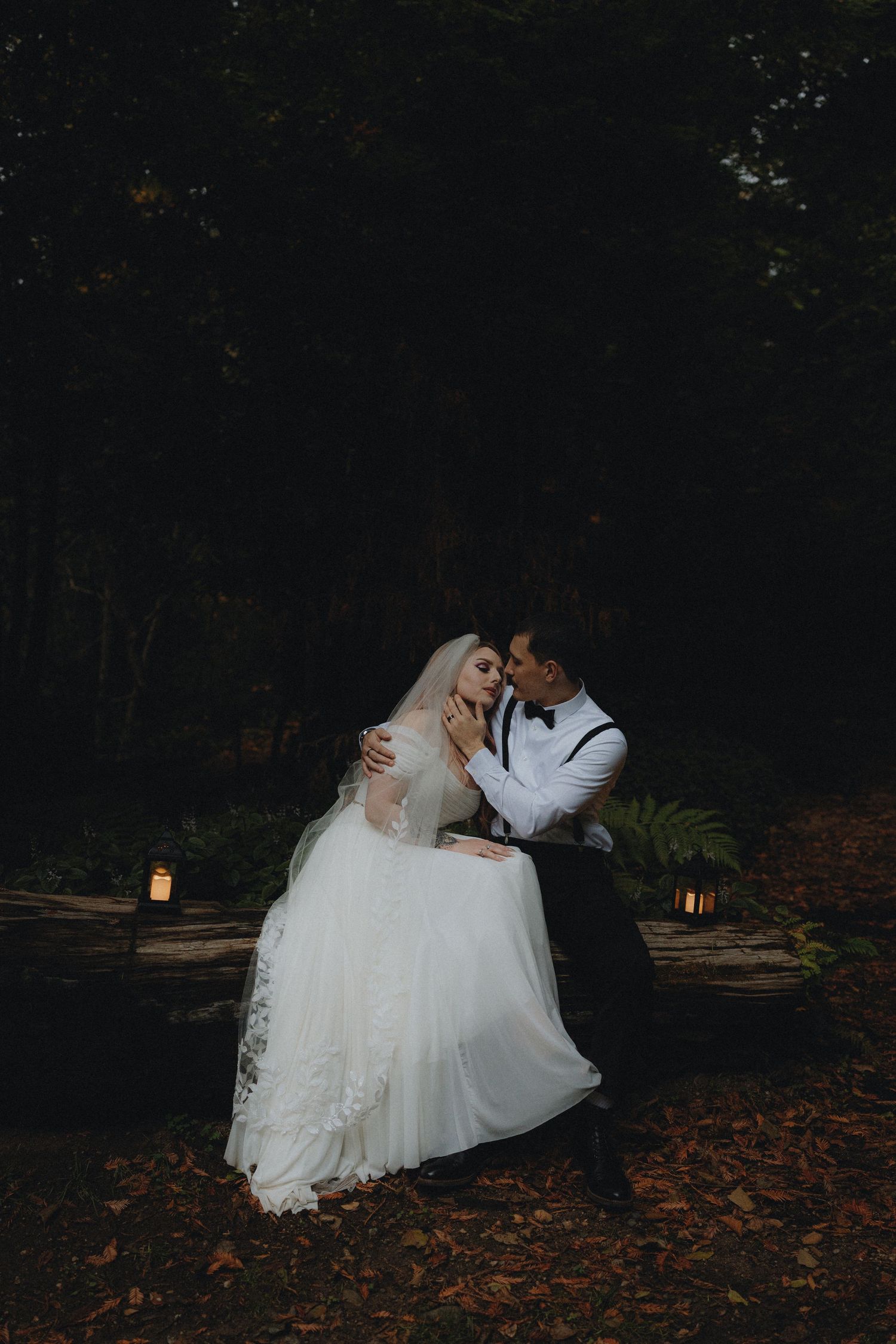 A romantic wedding couple dips in a dramatic pose on fallen autumn leaves at dusk with lanterns glowing in the background.