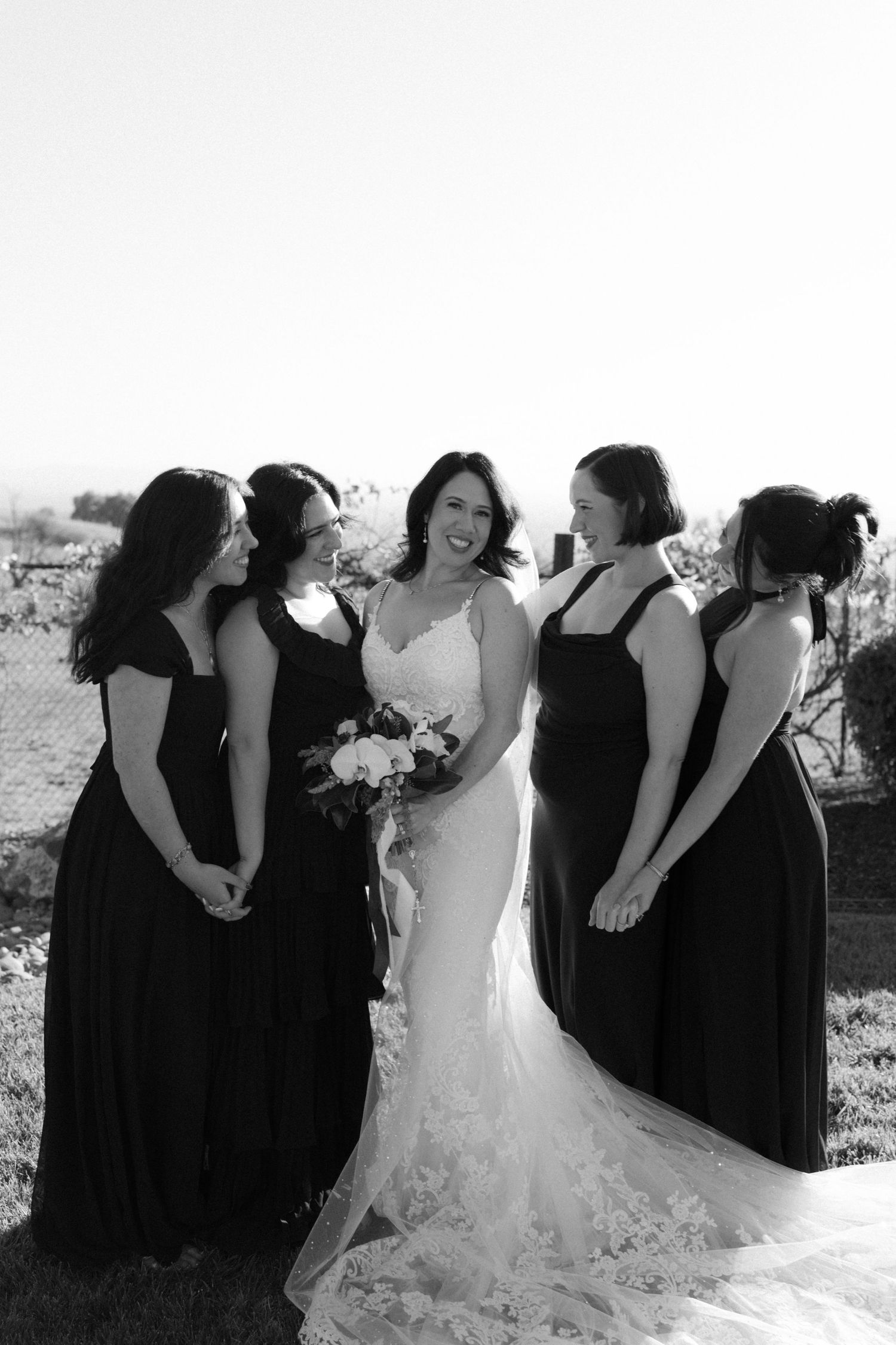 A bridal party in black dresses and white wedding gown pose together in natural sunlight.