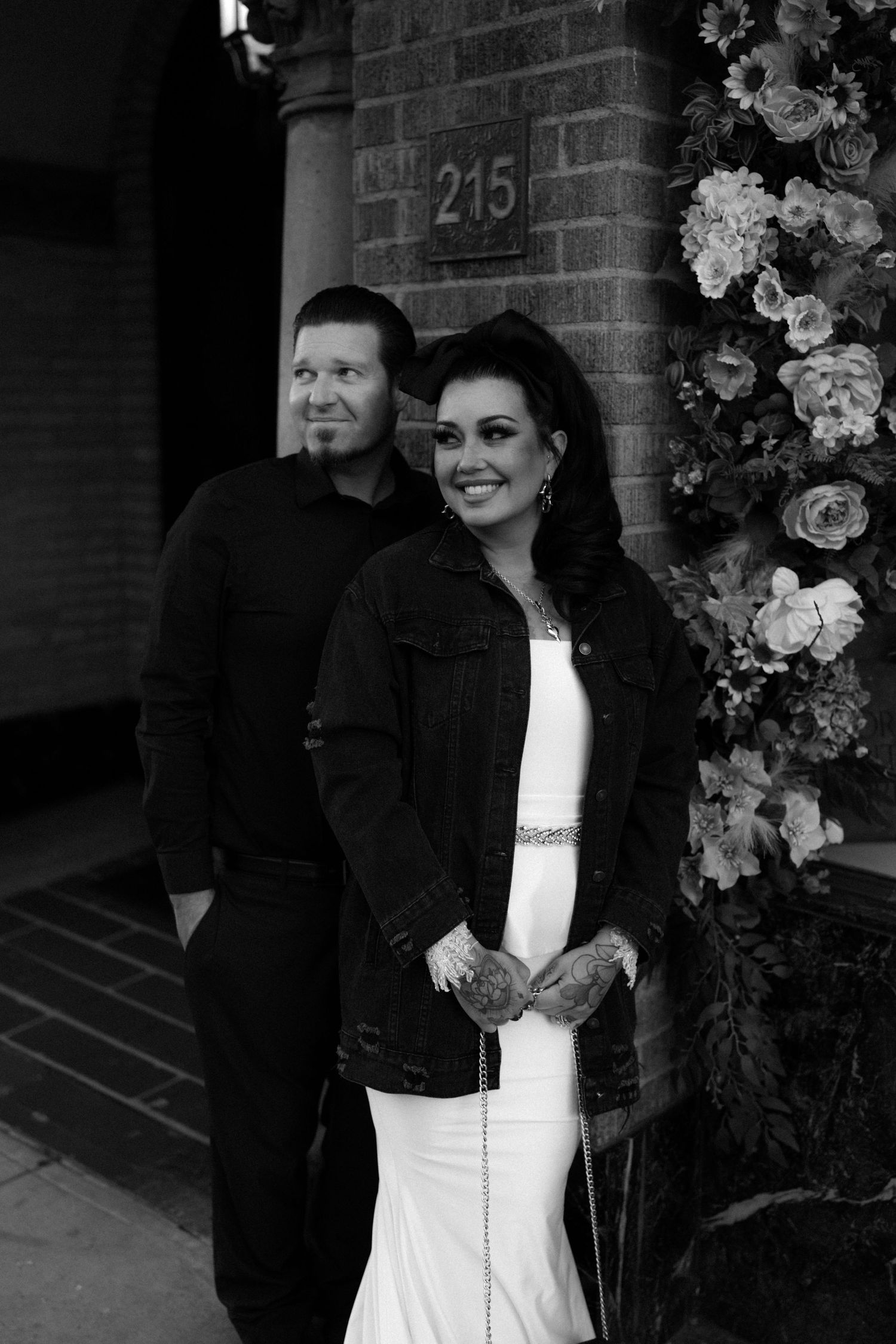 A wedding couple stands together near floral decorations and a brick wall.