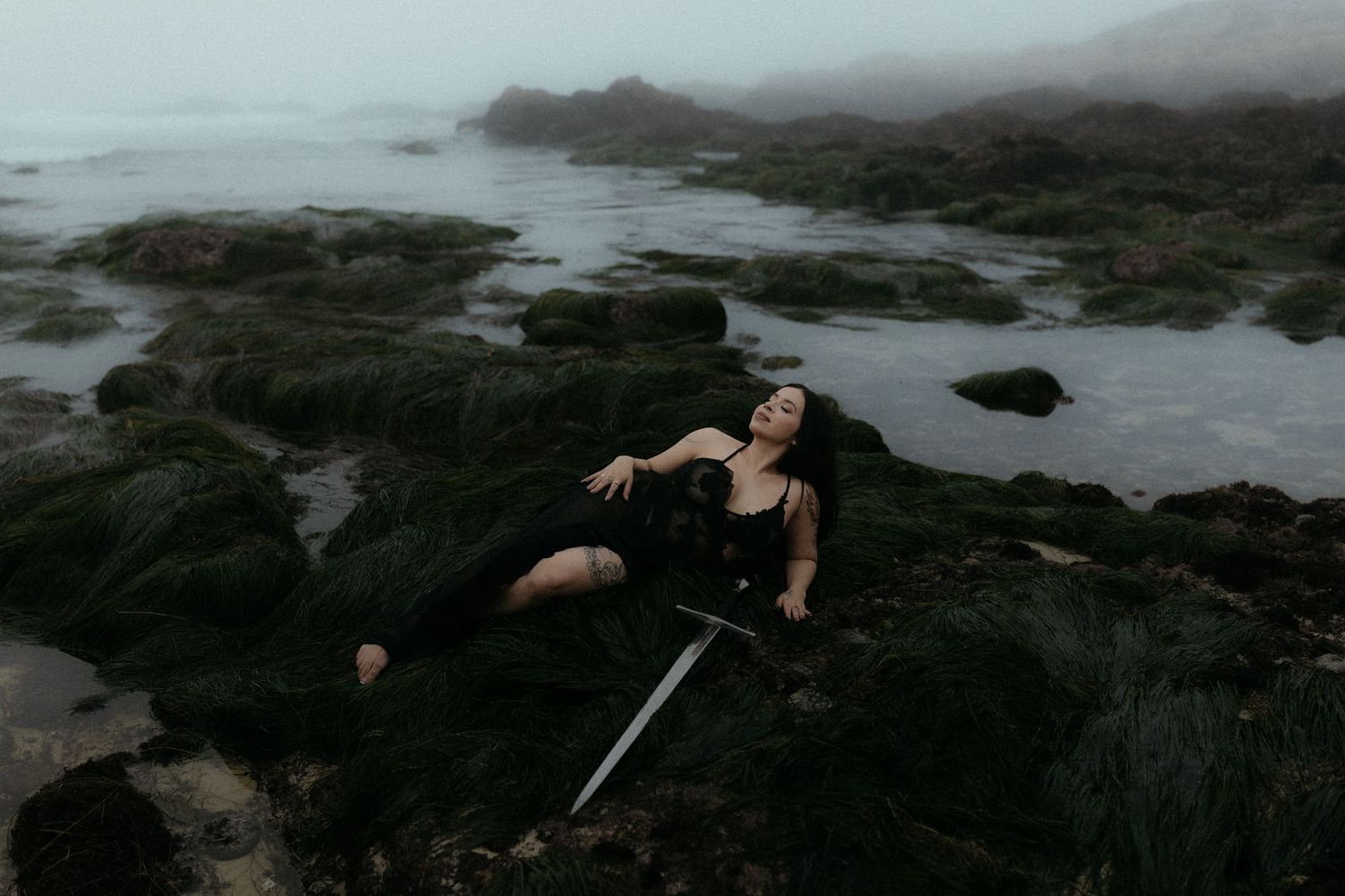 Dramatic coastal photoshoot featuring sword prop against misty ocean background.