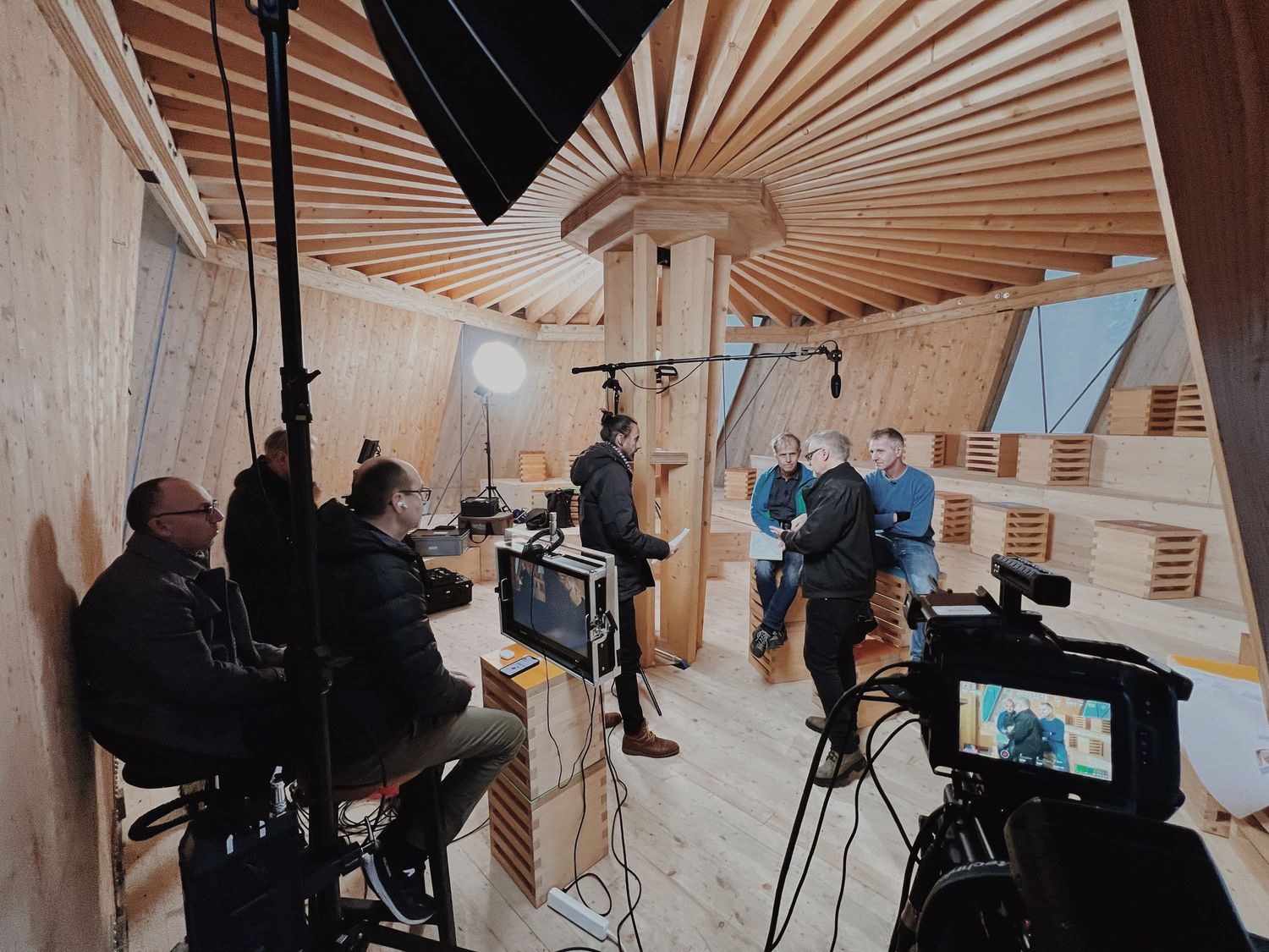 Film crew setting up equipment in a wooden room with fan-like ceiling details for a professional shoot.