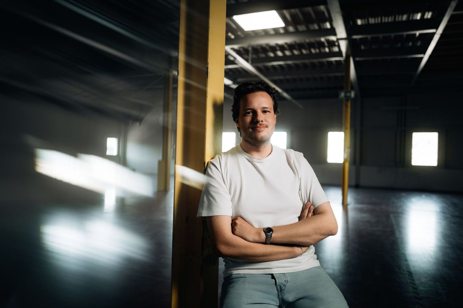 Man in white t-shirt stands confidently with arms crossed in a modern industrial space with dramatic lighting.