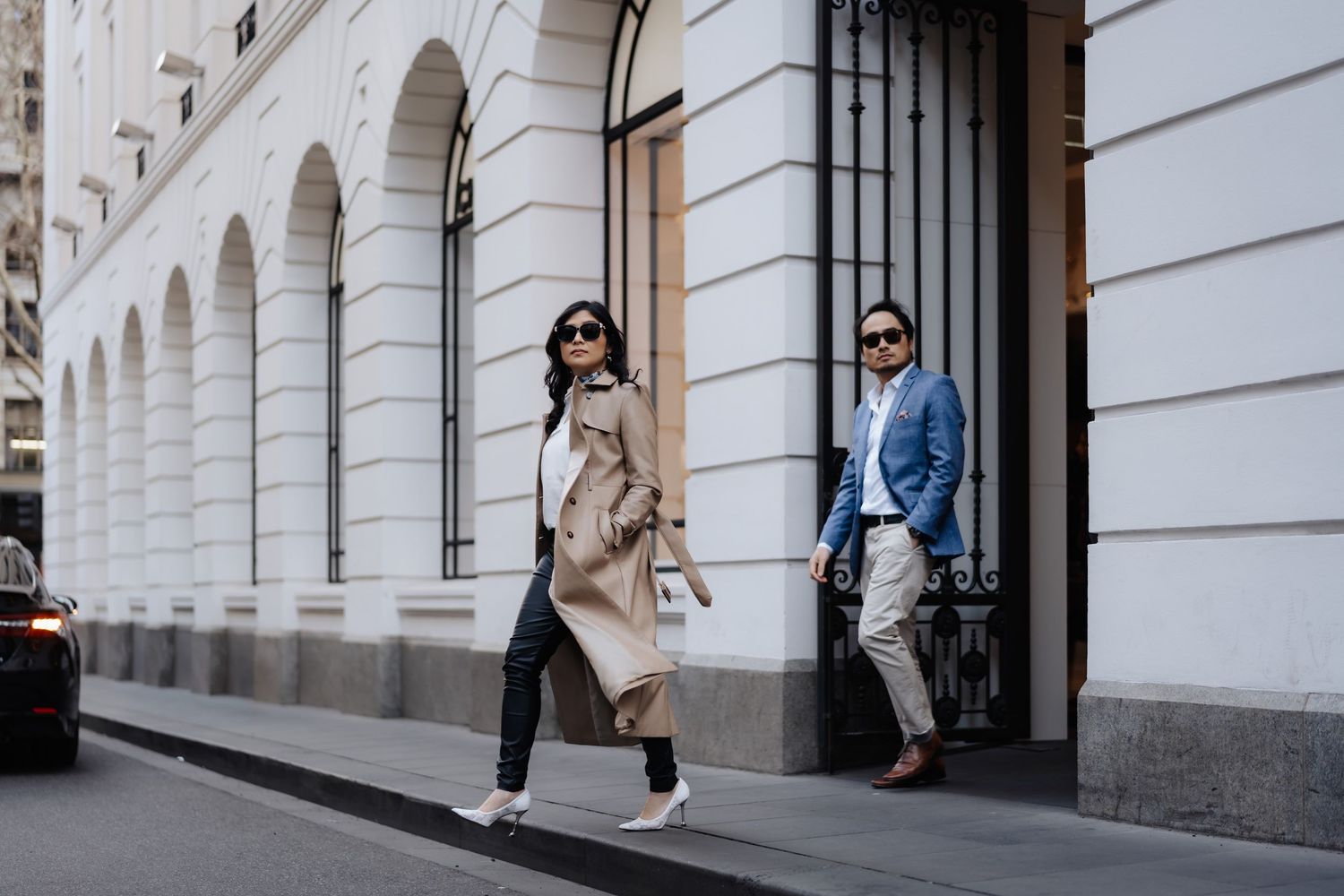 Stylish couple posing on a city street, woman in beige trench coat and man in blue blazer near white building.