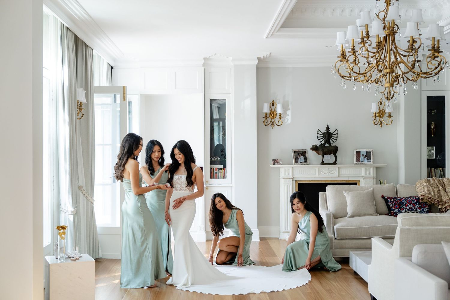Bride in white gown with bridesmaids in sage green dresses in elegant white room with chandelier.
