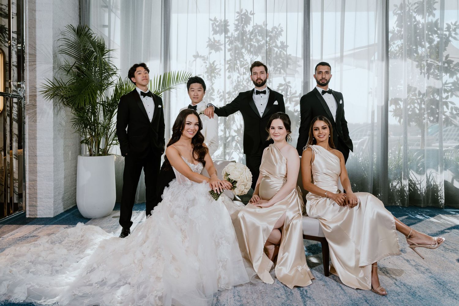 Wedding party portrait featuring bride in white gown, two bridesmaids in champagne dresses, and three groomsmen in black tuxedos.