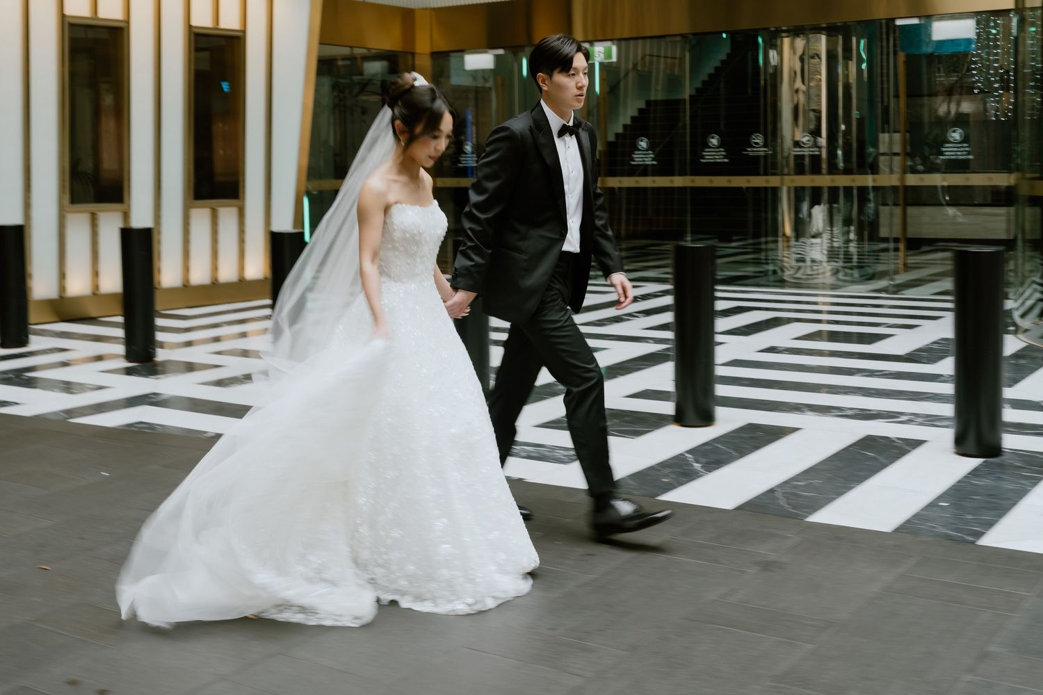 Asian bride in white ball gown and groom in black tuxedo walking together on black and white tiled floor.