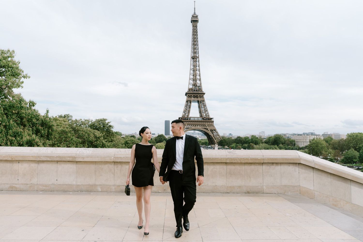 Couple in elegant black attire walking hand in hand at Trocadero with Eiffel Tower backdrop in Paris.