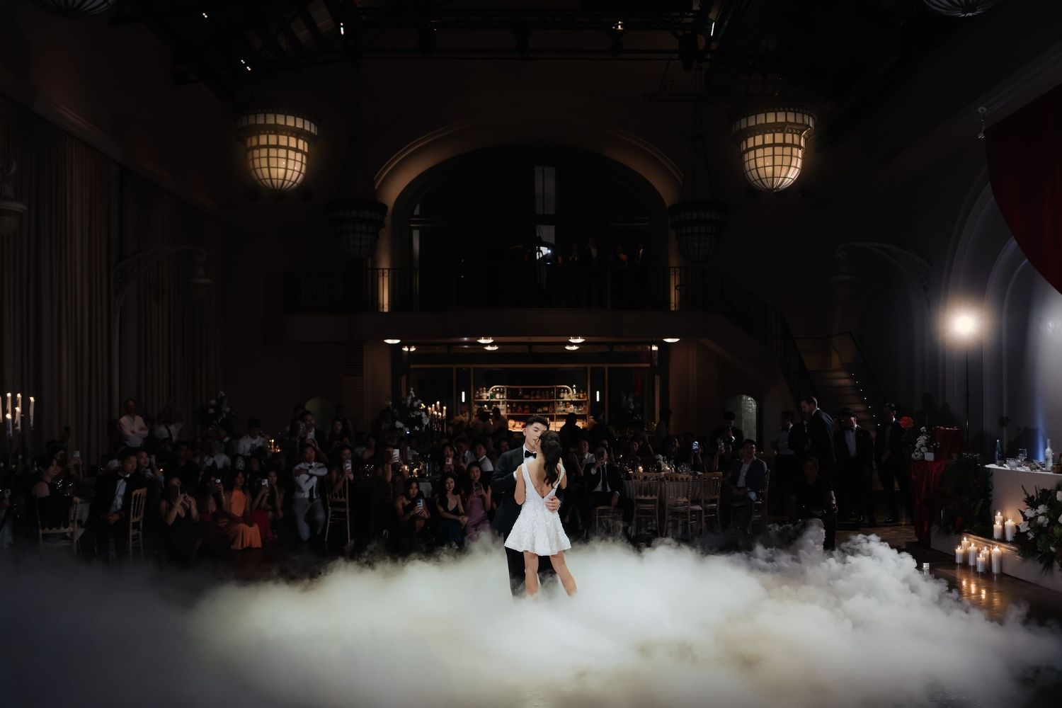 Bride and groom share first dance surrounded by dramatic fog machine clouds in elegant ballroom venue.