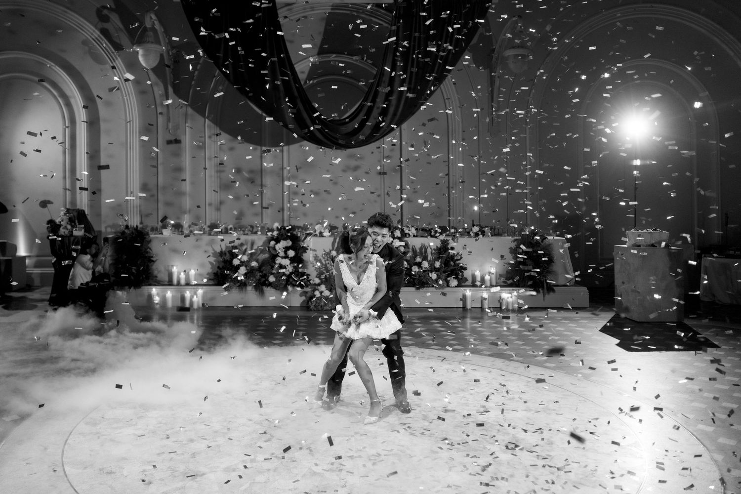Couple dancing on a white floor with confetti falling in an elegant ballroom during a wedding reception.