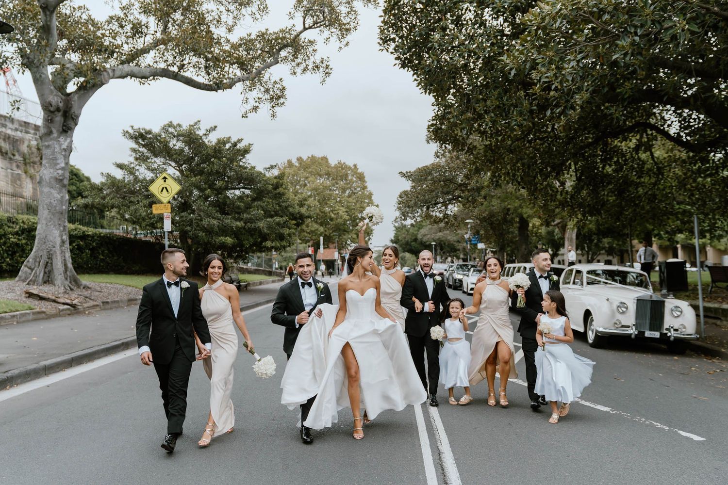 Wedding party crosses street in formal attire, bride in white gown with slit, groomsmen in tuxedos, bridesmaids in champagne dresses.