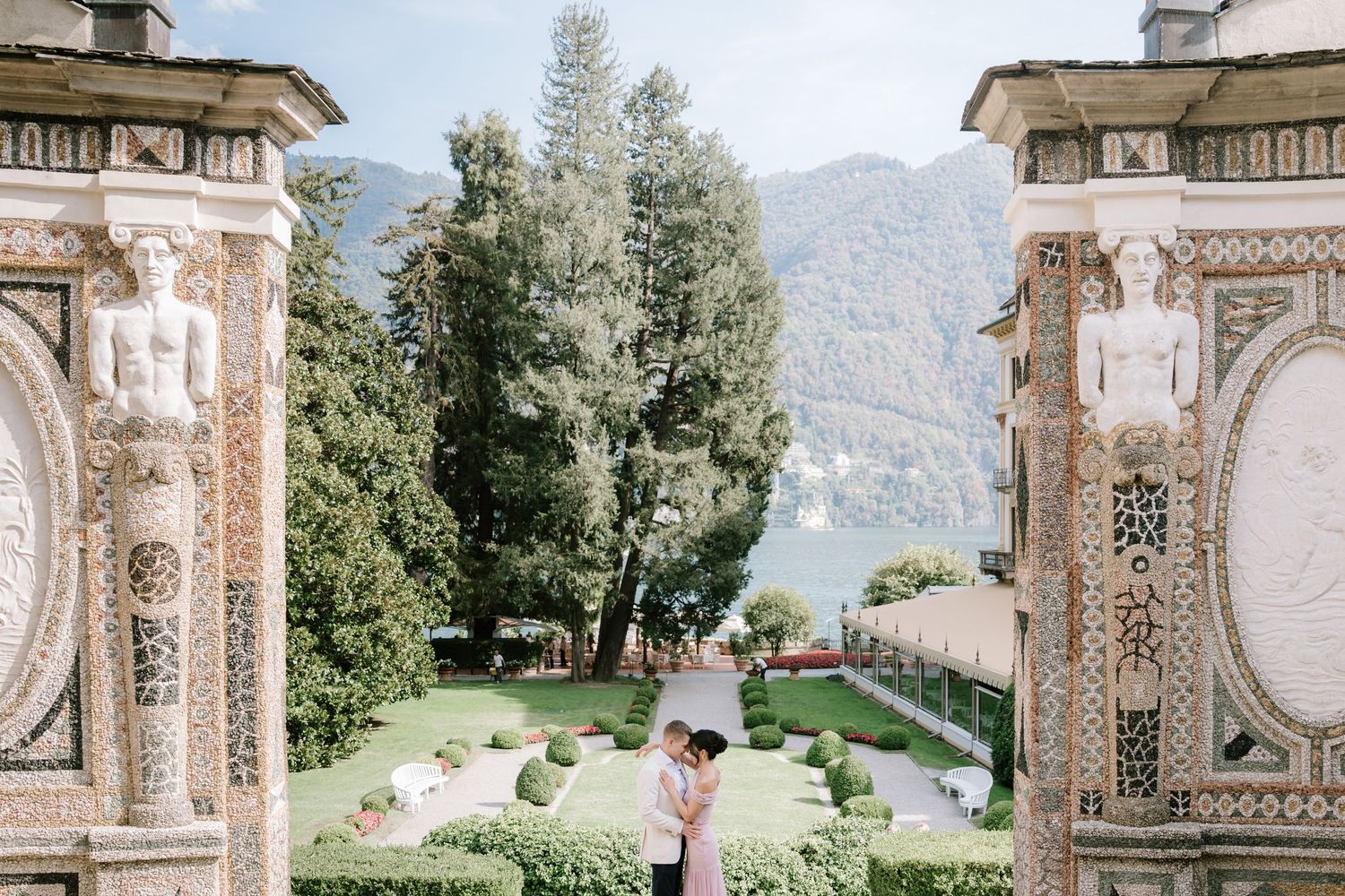 Couple embracing between ornate stone columns at Villa del Balbianello, Lake Como, with lush gardens and mountains behind.