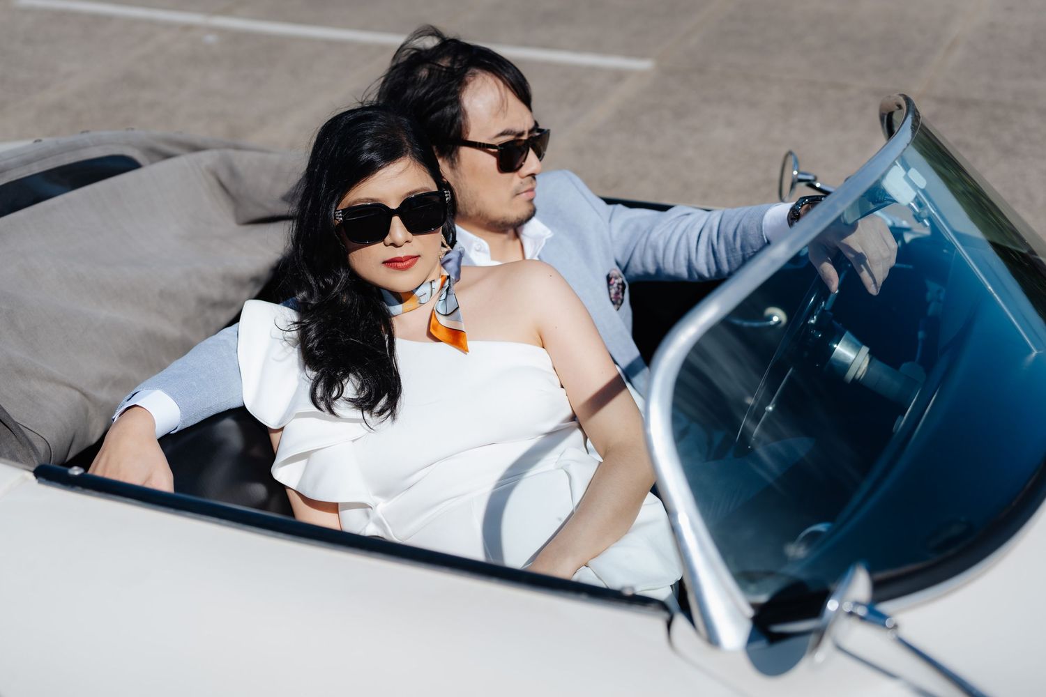 Stylish couple in sunglasses posing in a white convertible car, dressed in elegant white outfits.
