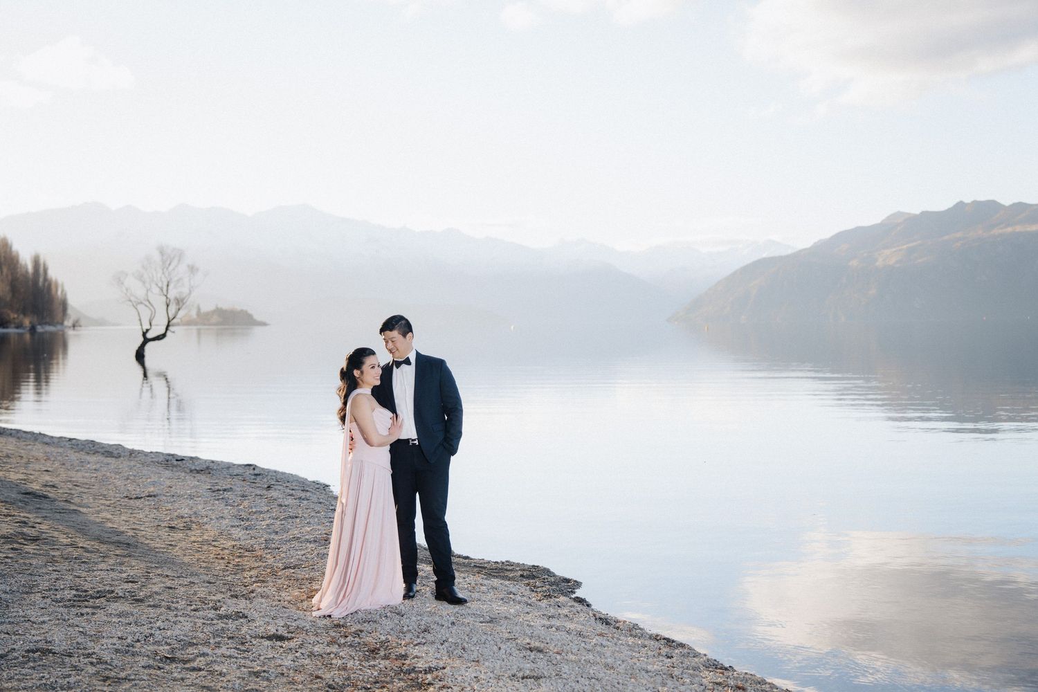Couple in formal attire poses by a serene lake with misty mountains and a lone tree reflected in calm waters. Two Peaches Wedding Photograph