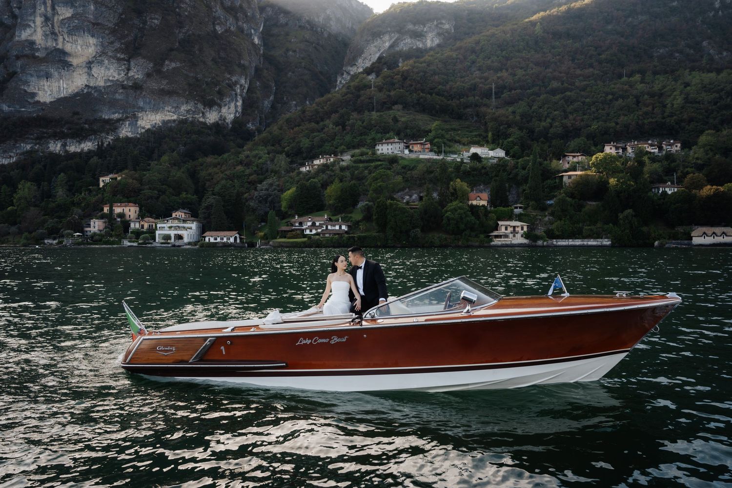 Couple posing on a classic wooden speedboat on a serene alpine lake with lush mountains in the background.