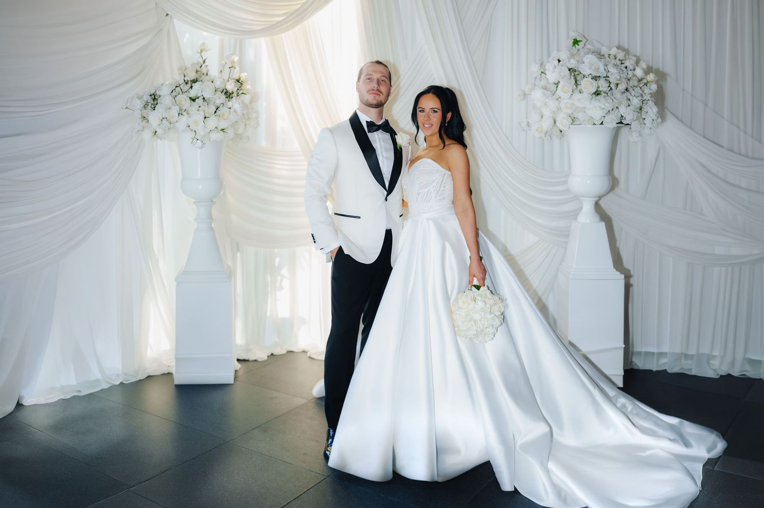 Elegant bride and groom posing before white floral backdrop, bride in strapless gown with train, groom in black tuxedo.