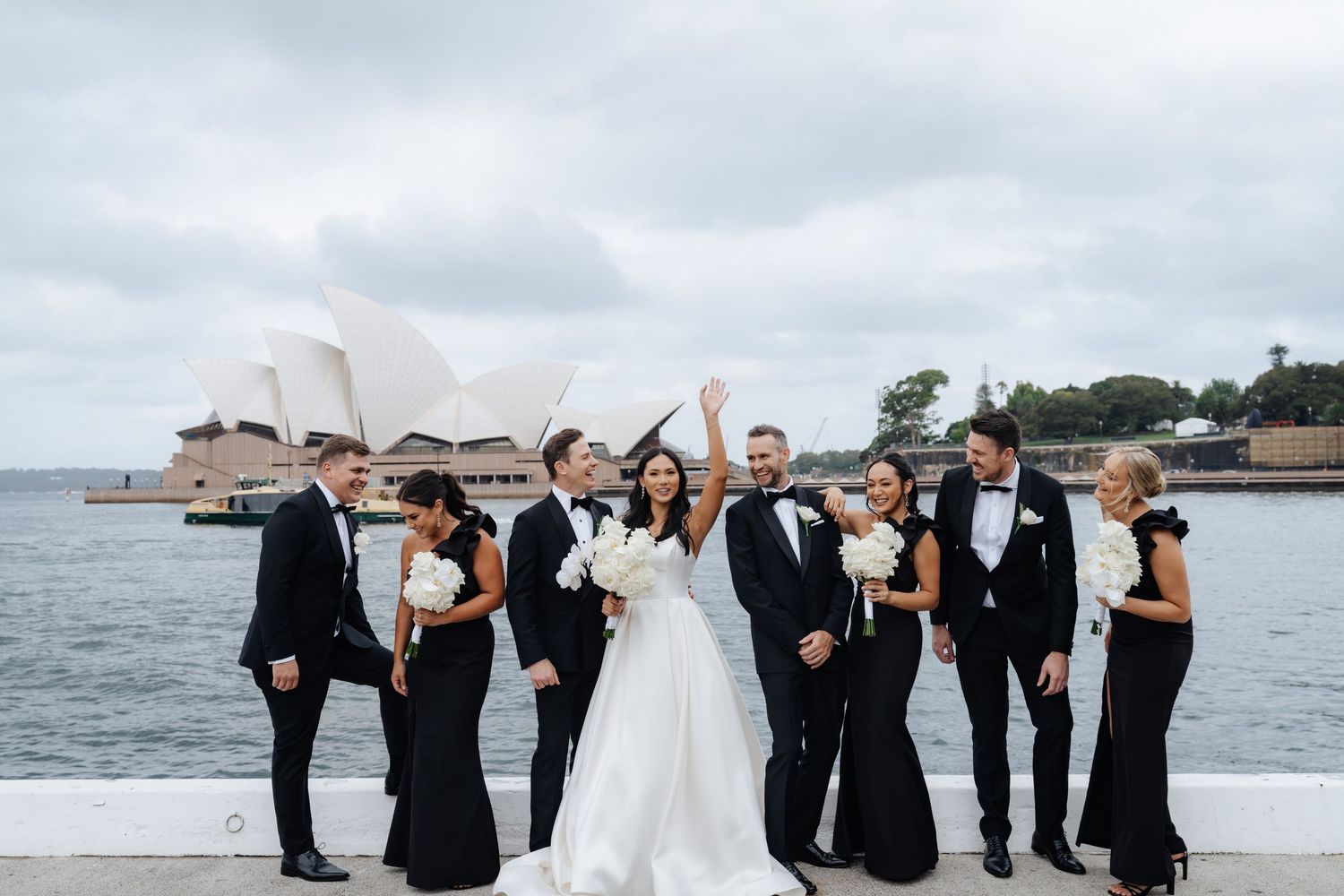 Wedding party in black and white attire celebrating in front of Sydney Opera House on a cloudy day.
