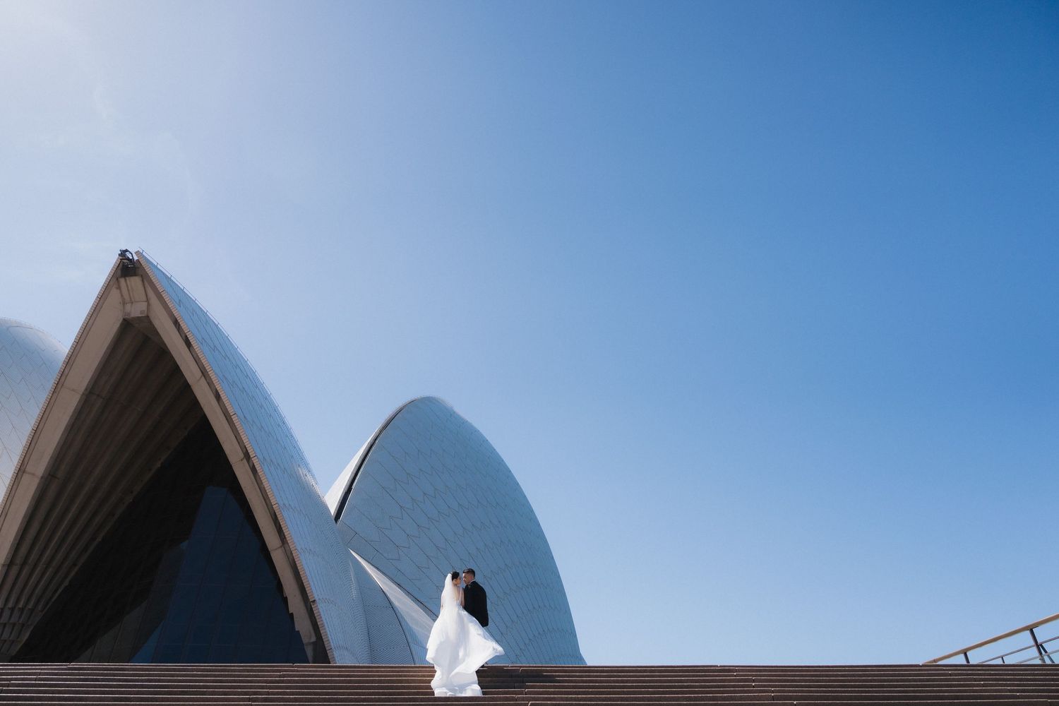 Couple in wedding attire standing on steps of Sydney Opera House under clear blue sky.