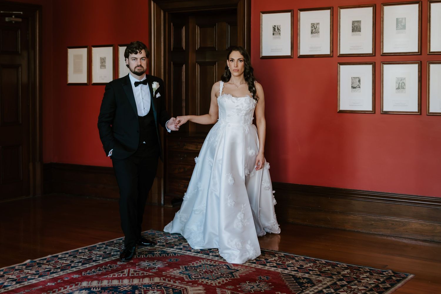 Elegant bride and groom holding hands in a red-walled gallery room, wearing a white ball gown and black tuxedo.
