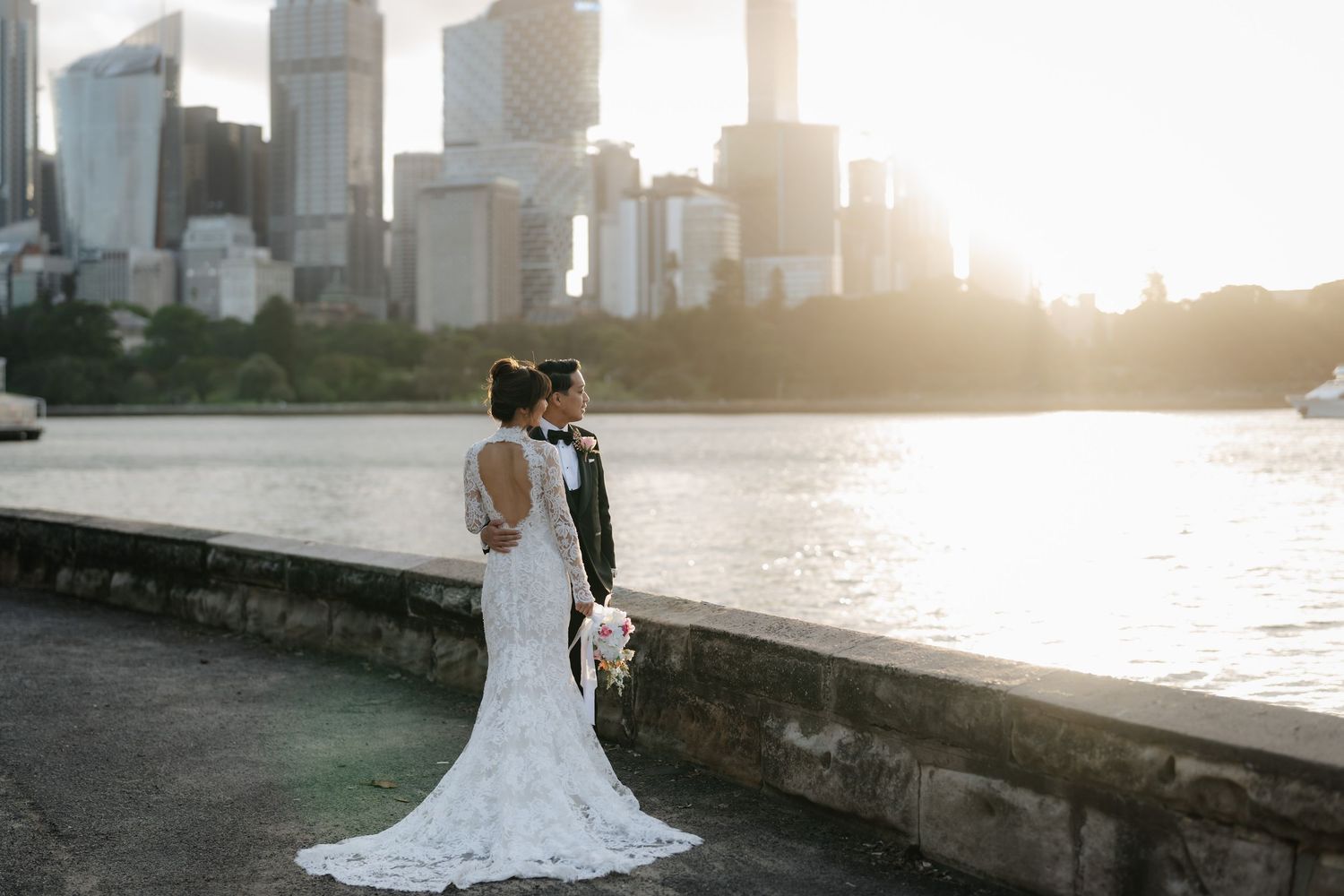 Bride and groom pose by waterfront with stunning city skyline backdrop during golden hour sunset.