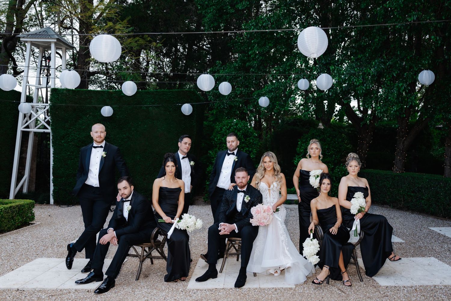 Wedding party in black tie attire poses outdoors with white lanterns and lush greenery backdrop.