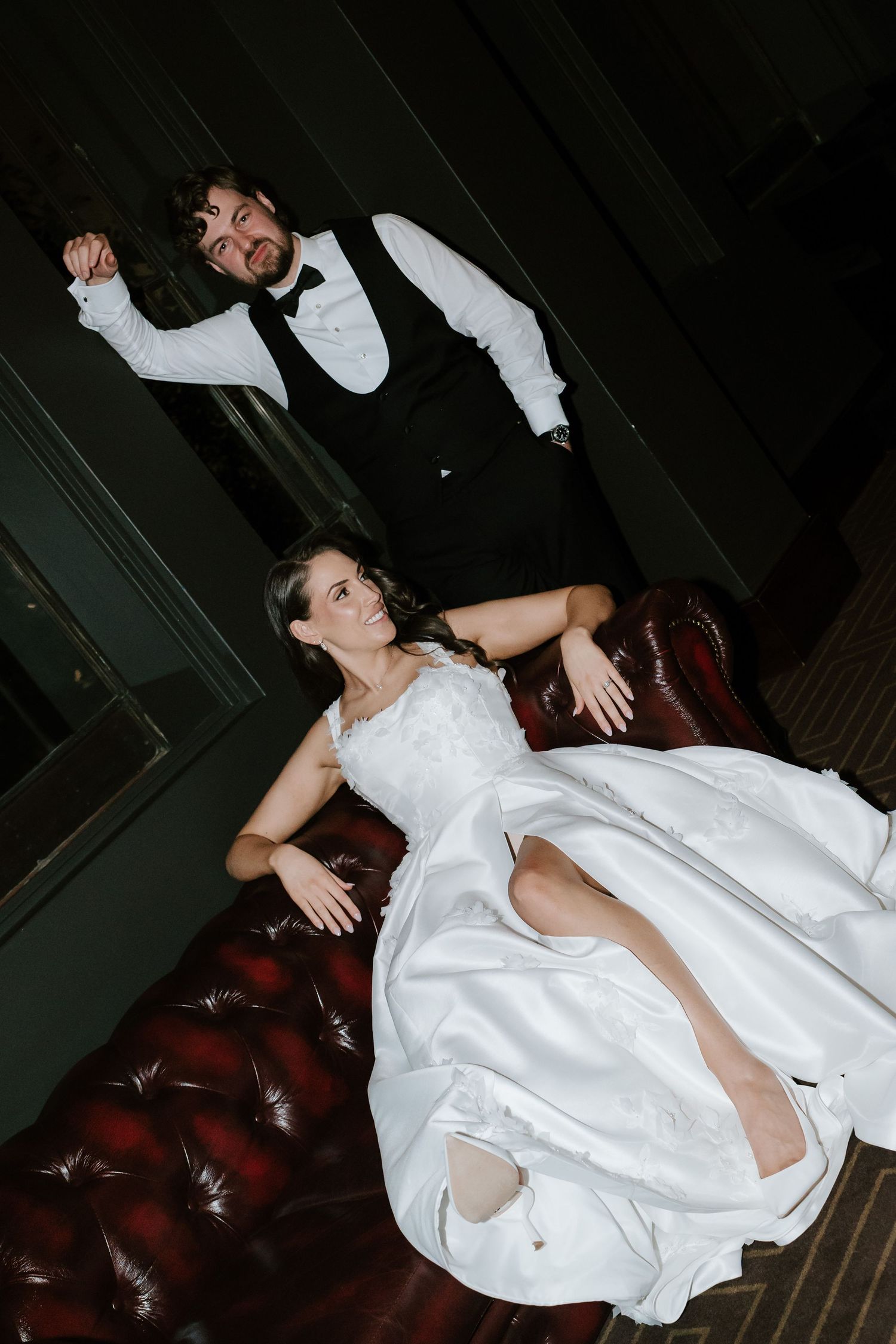 Bride in white gown reclines on leather sofa while groom in vest and bow tie celebrates behind her at wedding reception.