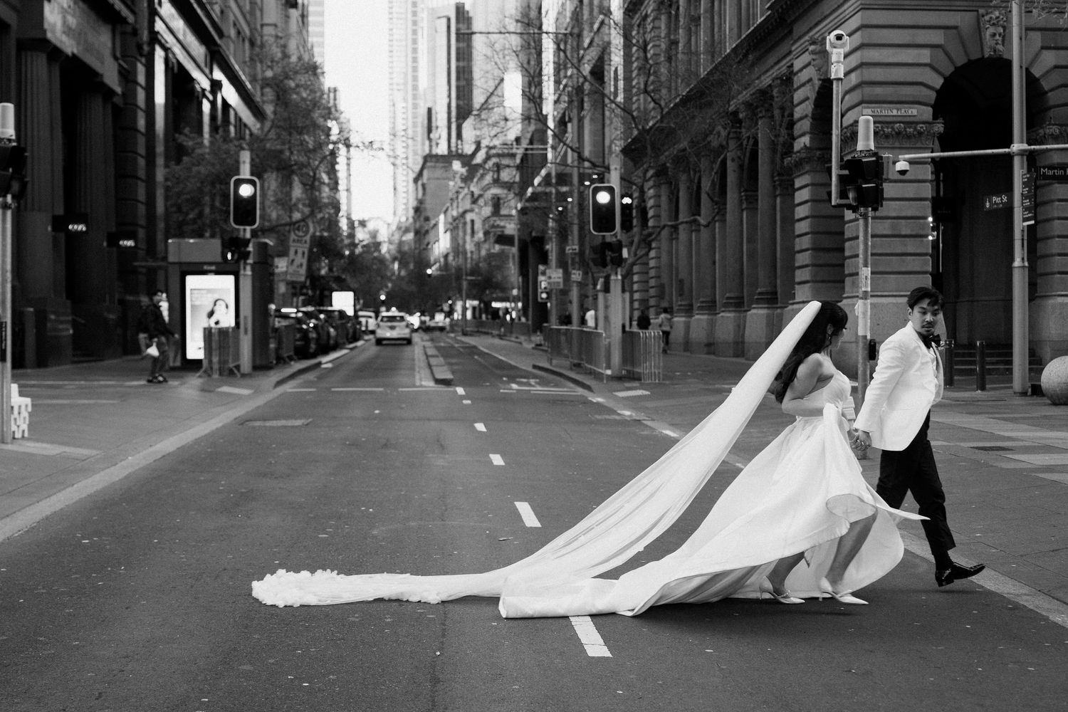 Black and white photo of bride and groom crossing a city street, her long veil flowing dramatically in the wind.