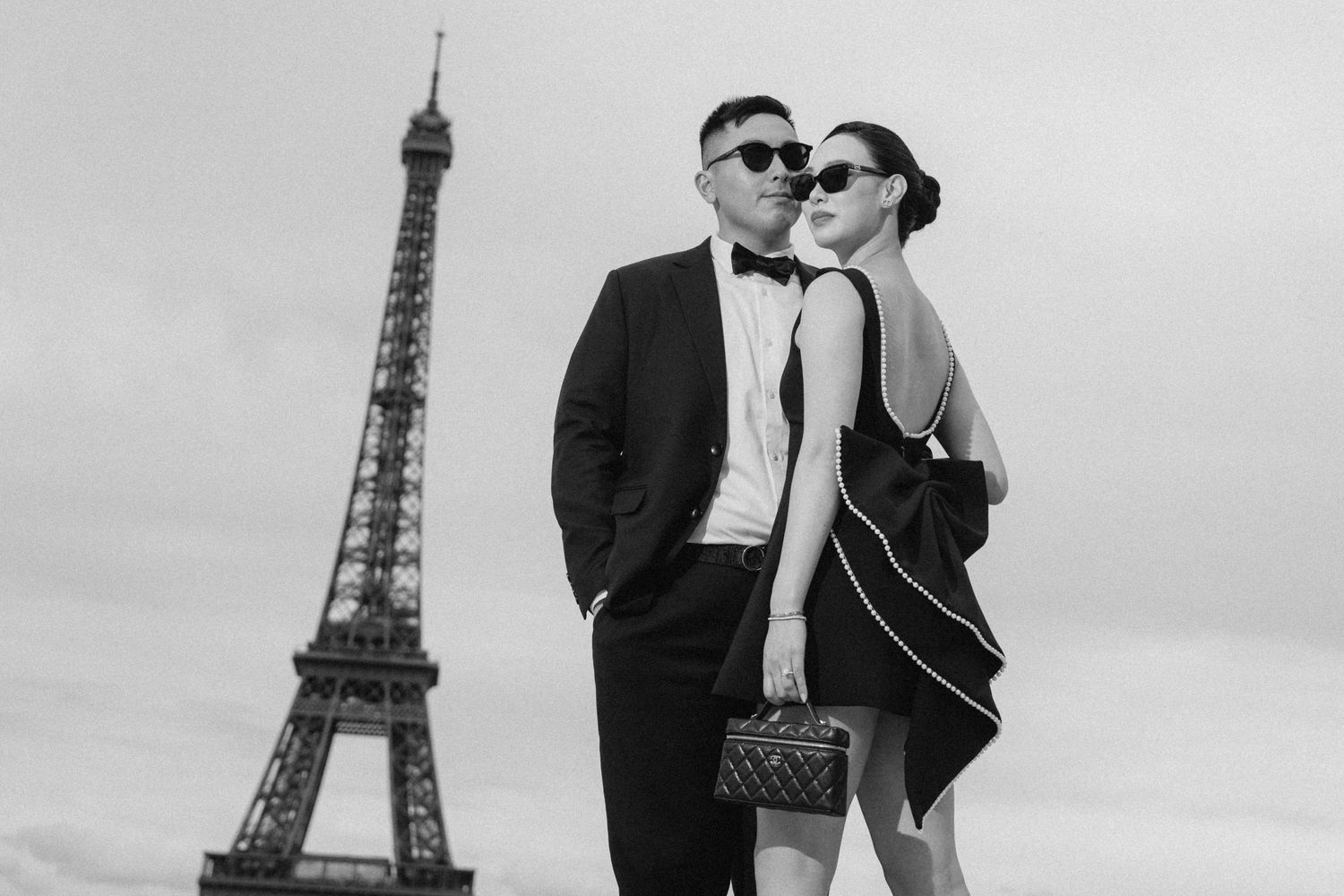 Elegant couple posing in front of the Eiffel Tower in Paris, wearing formal black attire in a stylish black and white photo.