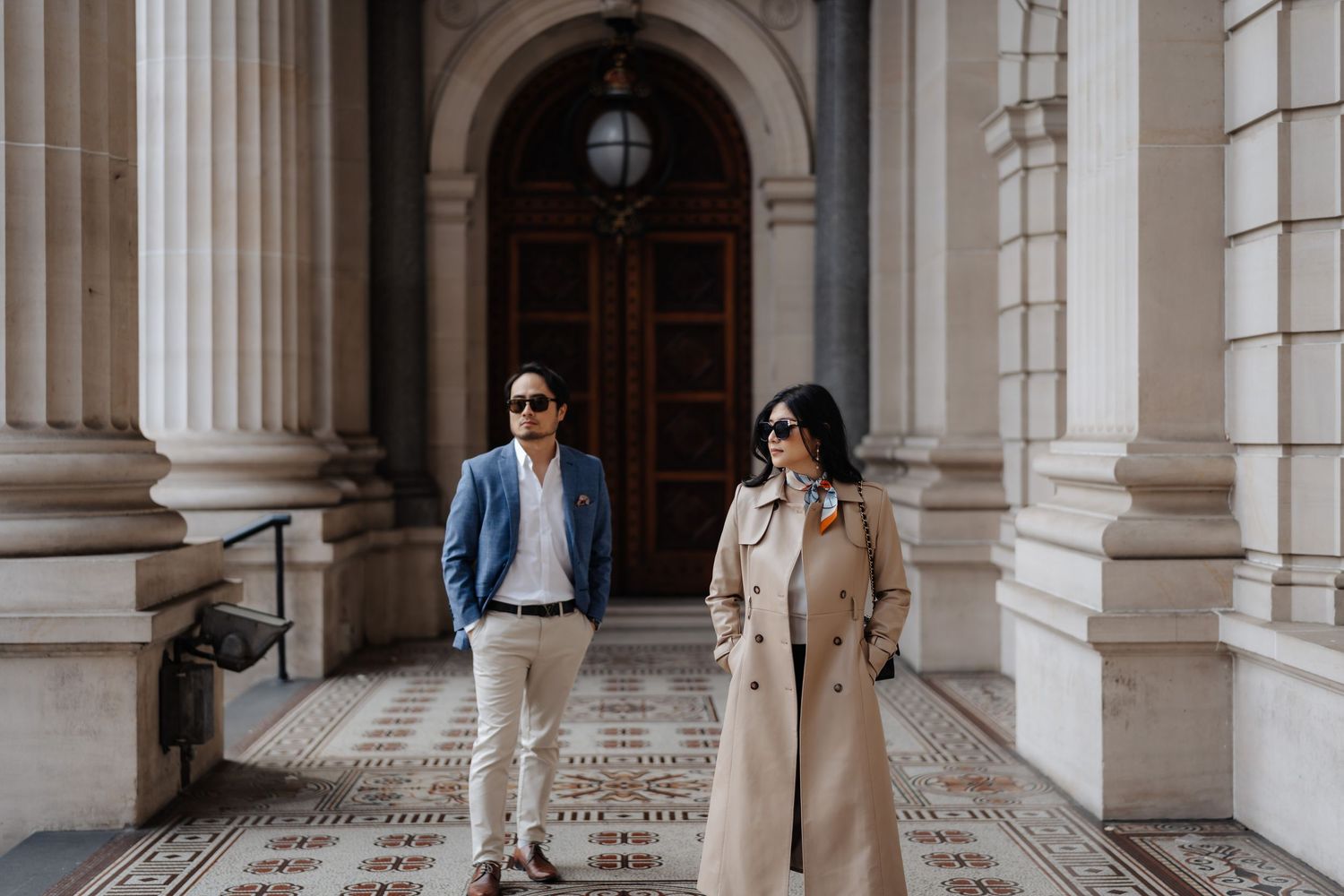 Couple posing outside a grand building with ornate columns and arched doorway on a tiled walkway.