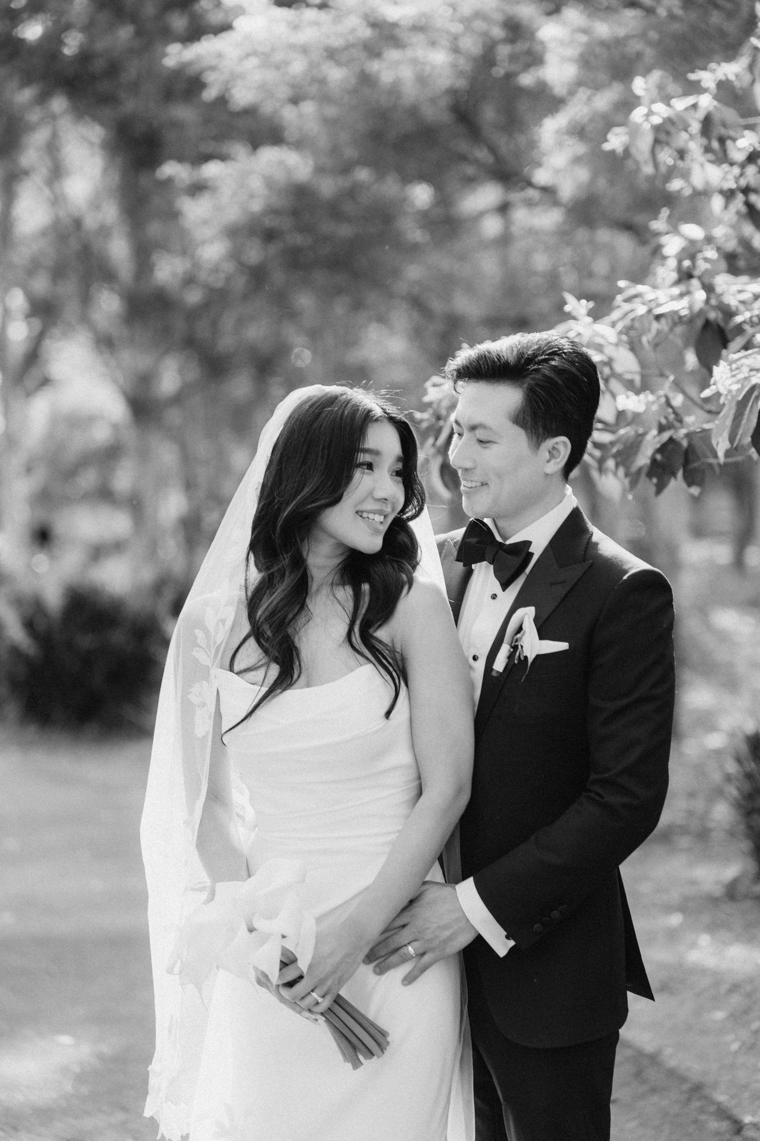 Black and white wedding portrait of a smiling bride in a white gown and groom in a tuxedo embracing outdoors.