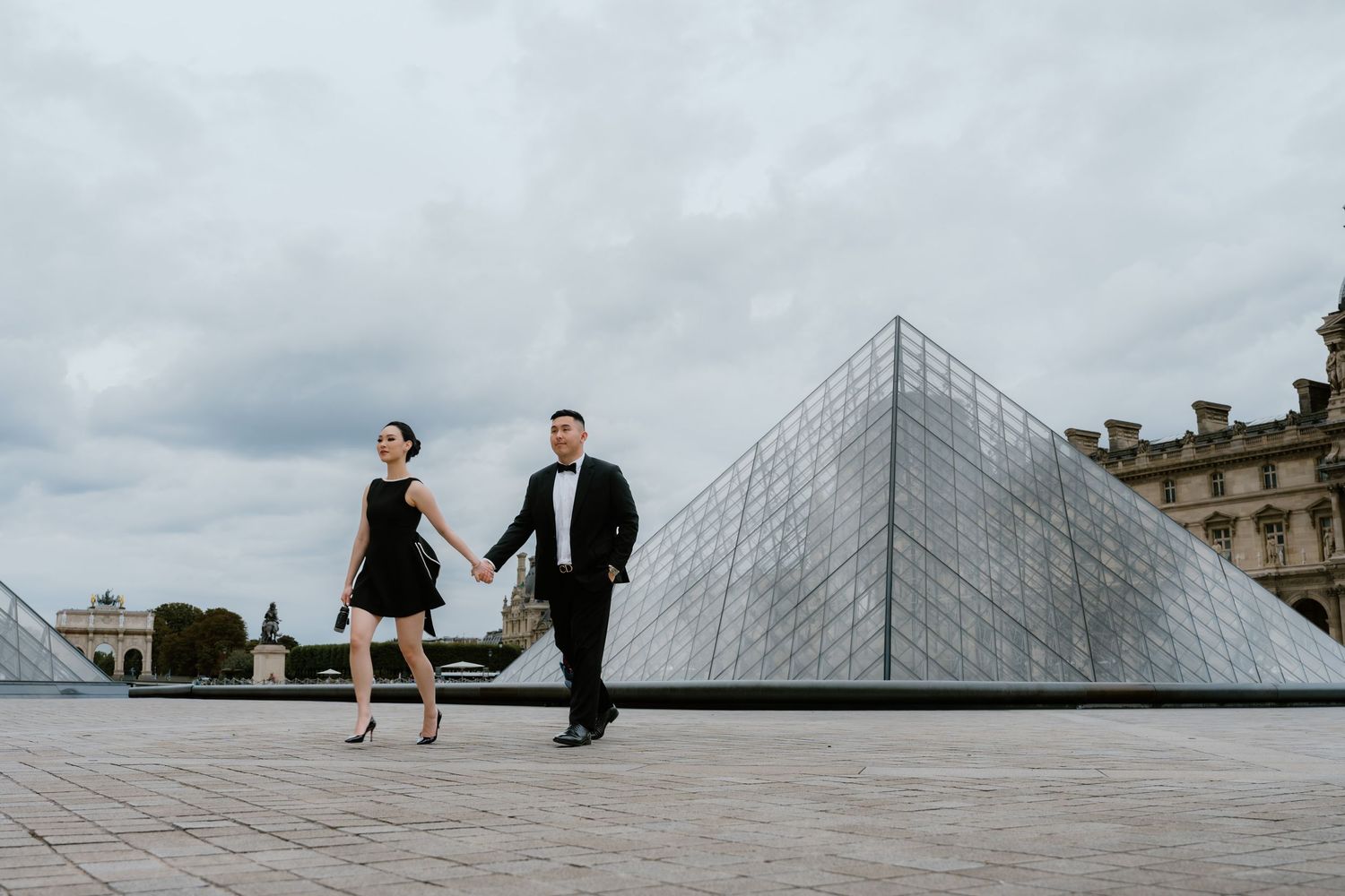 Couple holding hands walking in front of the Louvre Pyramid in Paris, woman in black dress, man in tuxedo.