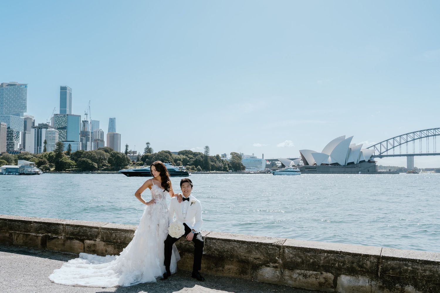 Bride and groom posing by Sydney Harbour with Opera House and Harbour Bridge in background on sunny wedding day.